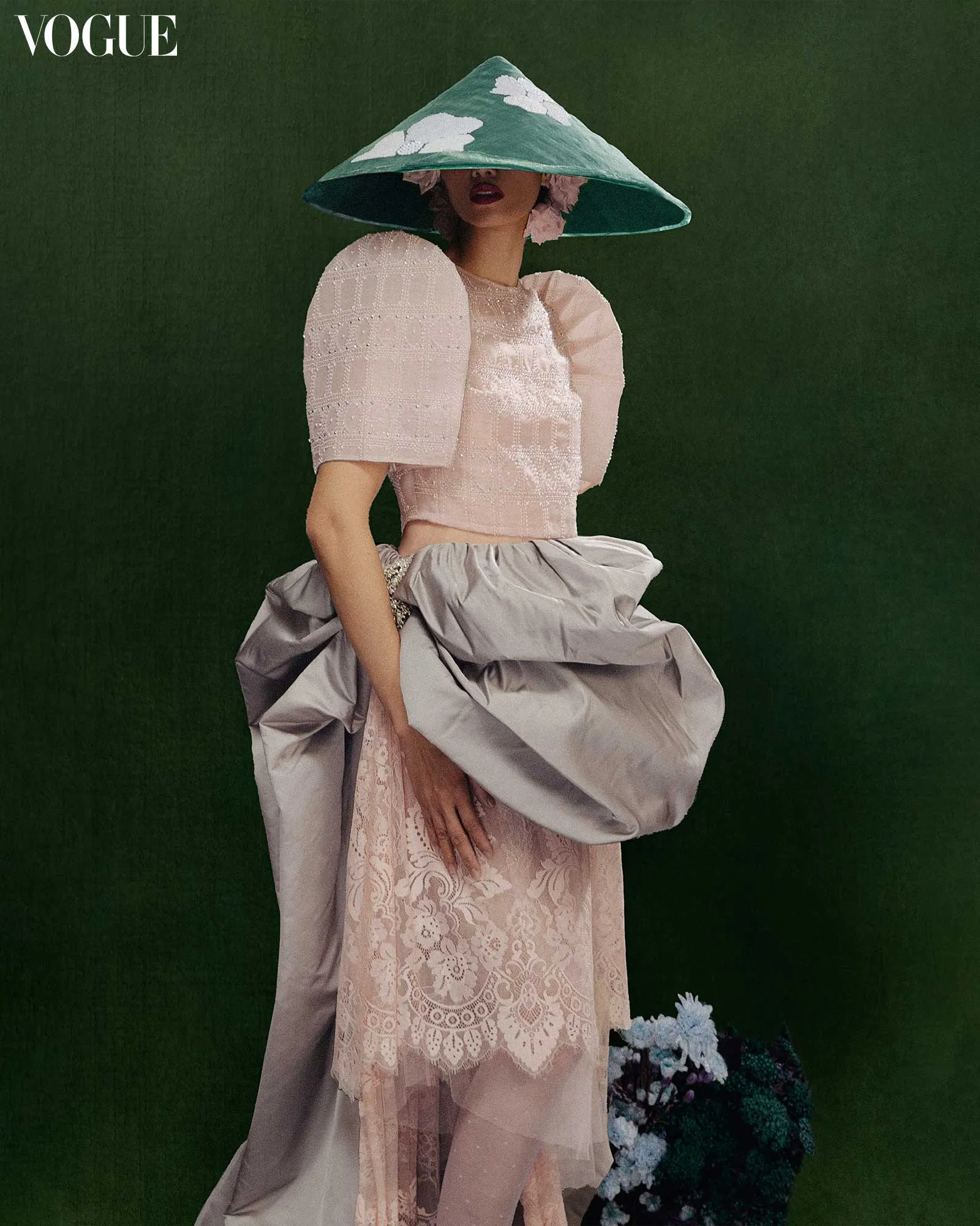 Model in pale pink lace dress with oversized sleeves and a teal conical hat against a dark green backdrop; Vogue logo top-left.