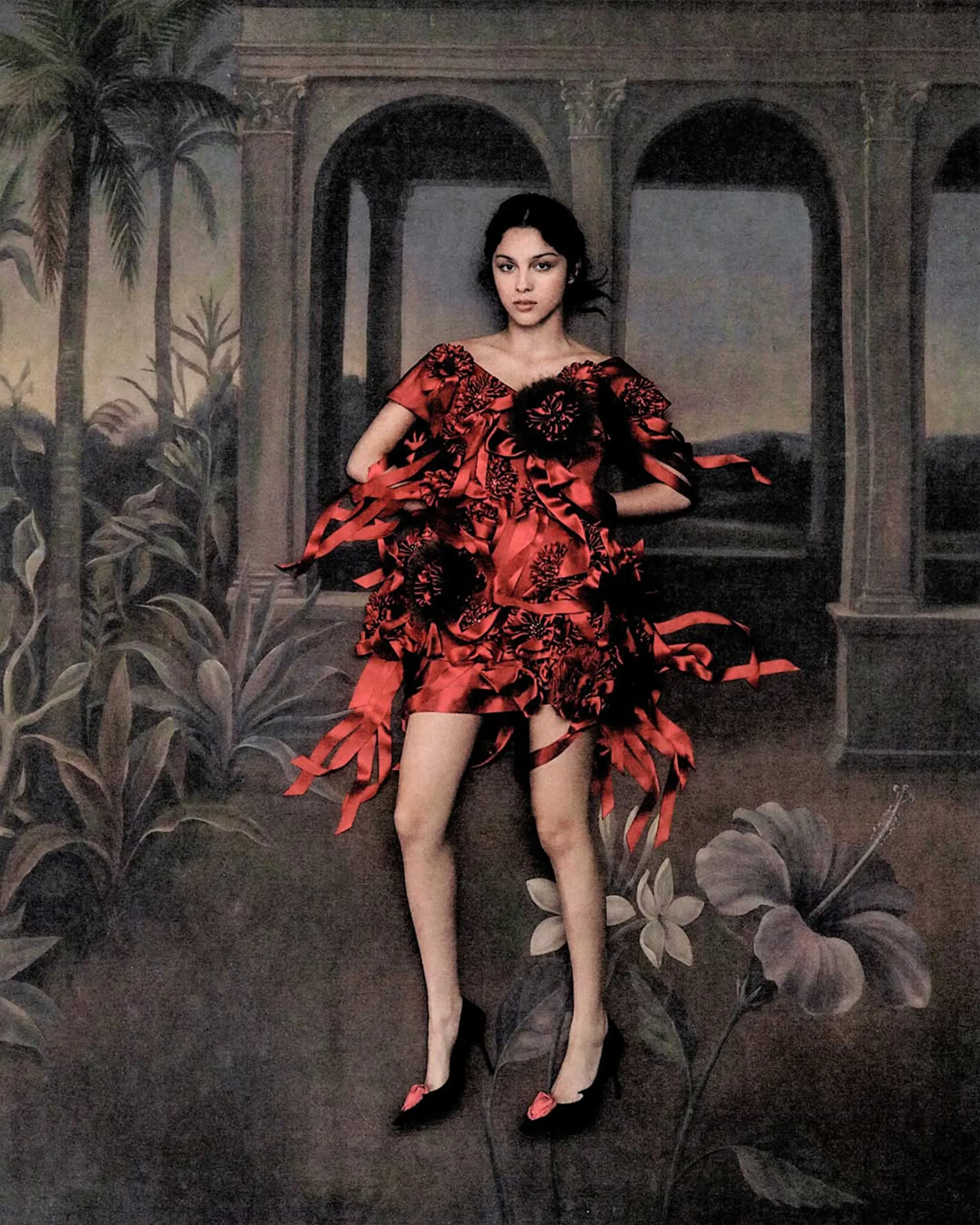 Woman in a red floral dress poses with hands on hips in front of a painted archway and tropical plants.