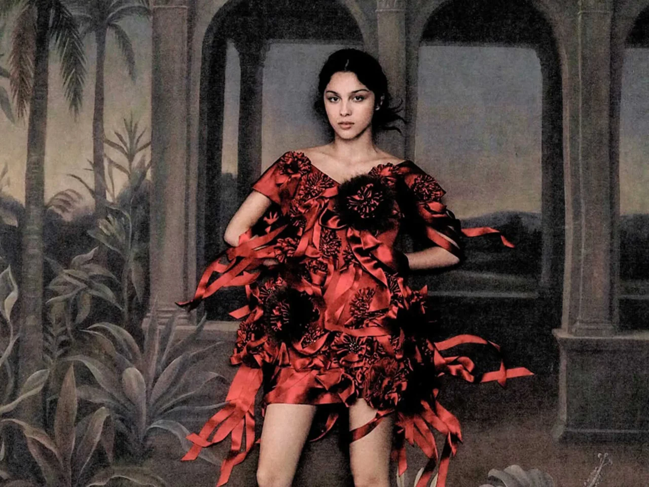 Woman in a dramatic red off-shoulder dress with black floral patterns, posing with hands on hips against a tropical backdrop.