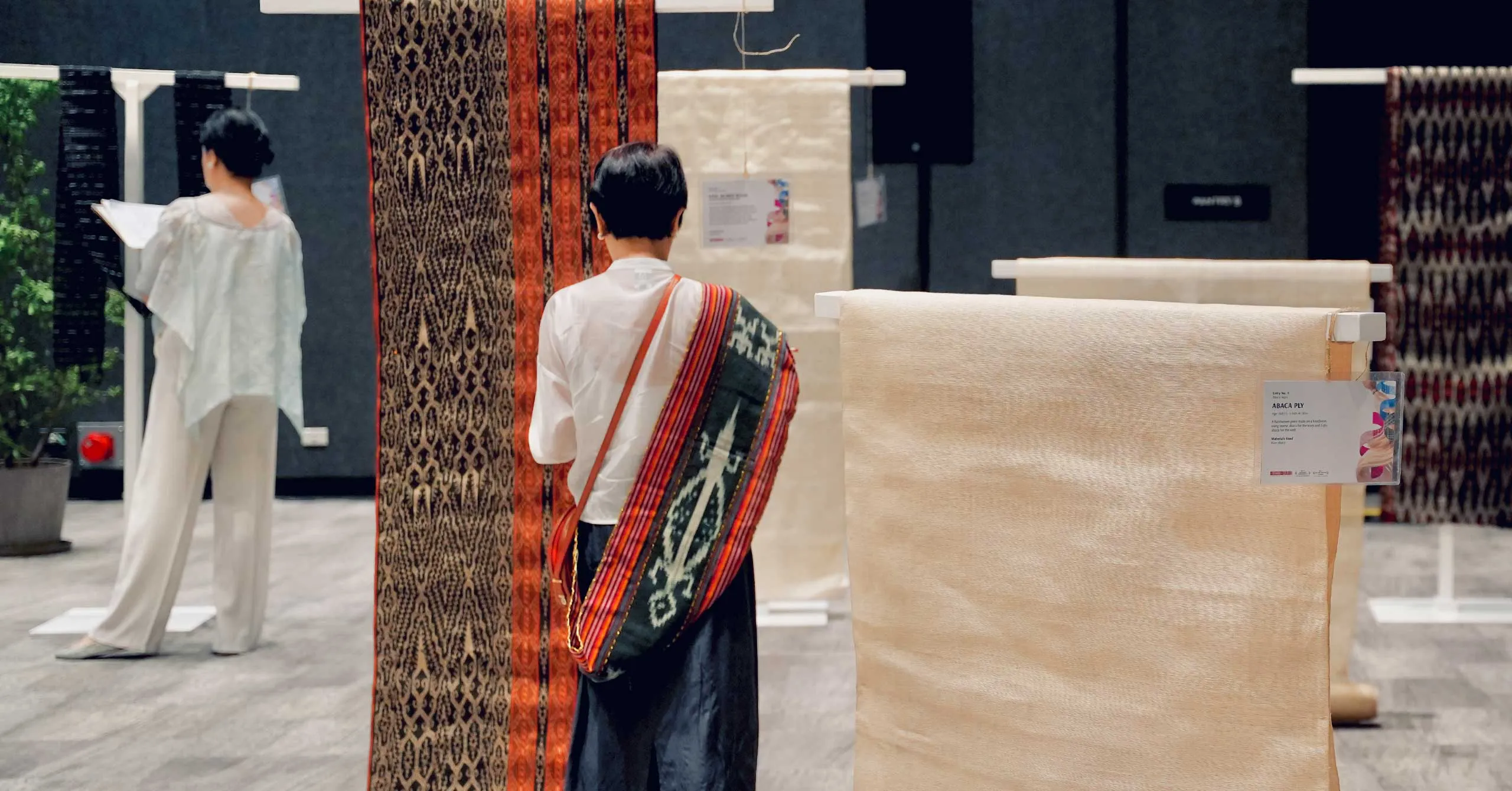 Person wearing a colorful woven shawl draped over the shoulder examines fabric rolls in a textile showroom, with hanging patterns behind them.