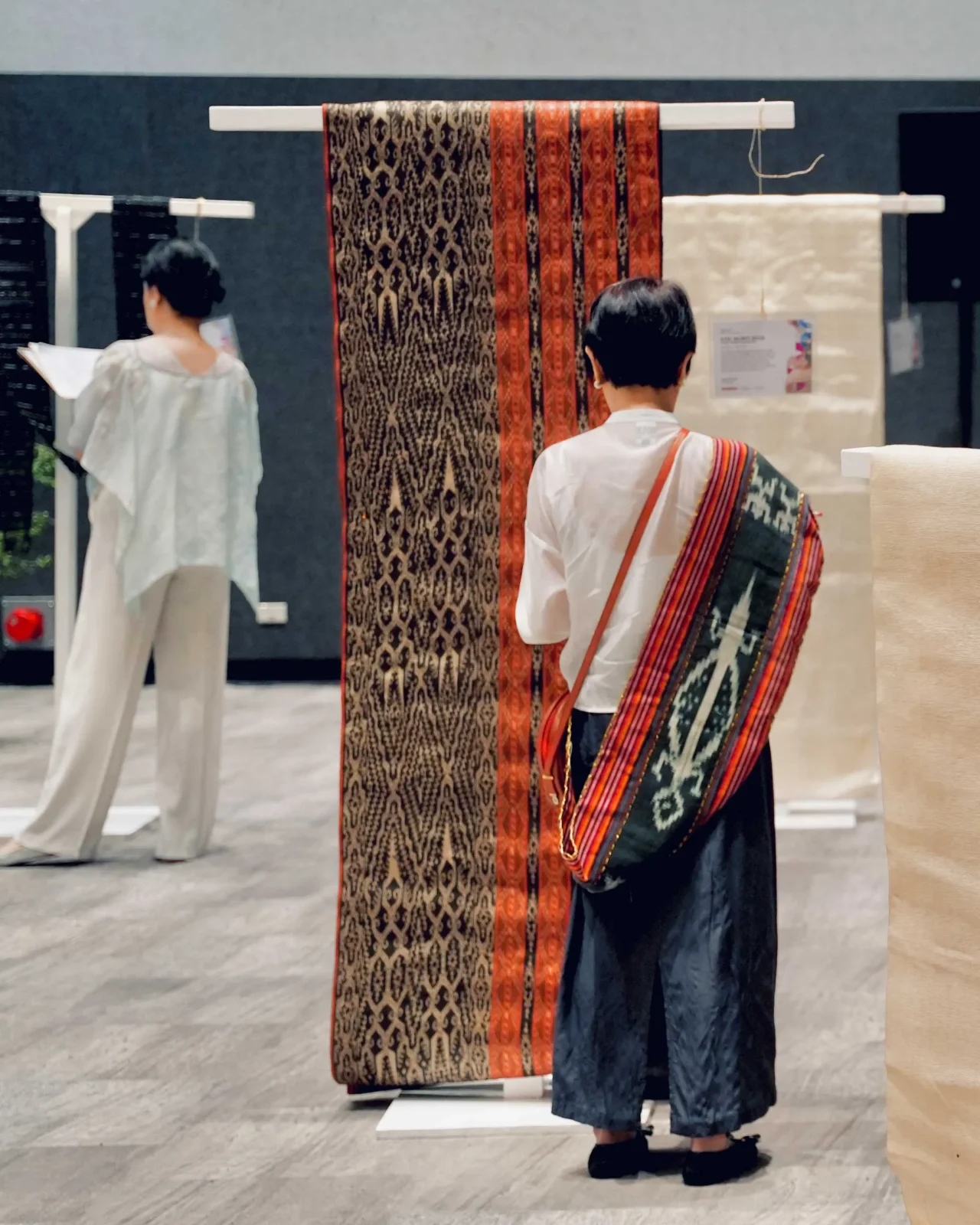 Person wearing a colorful woven shawl draped over the shoulder examines fabric rolls in a textile showroom, with hanging patterns behind them.