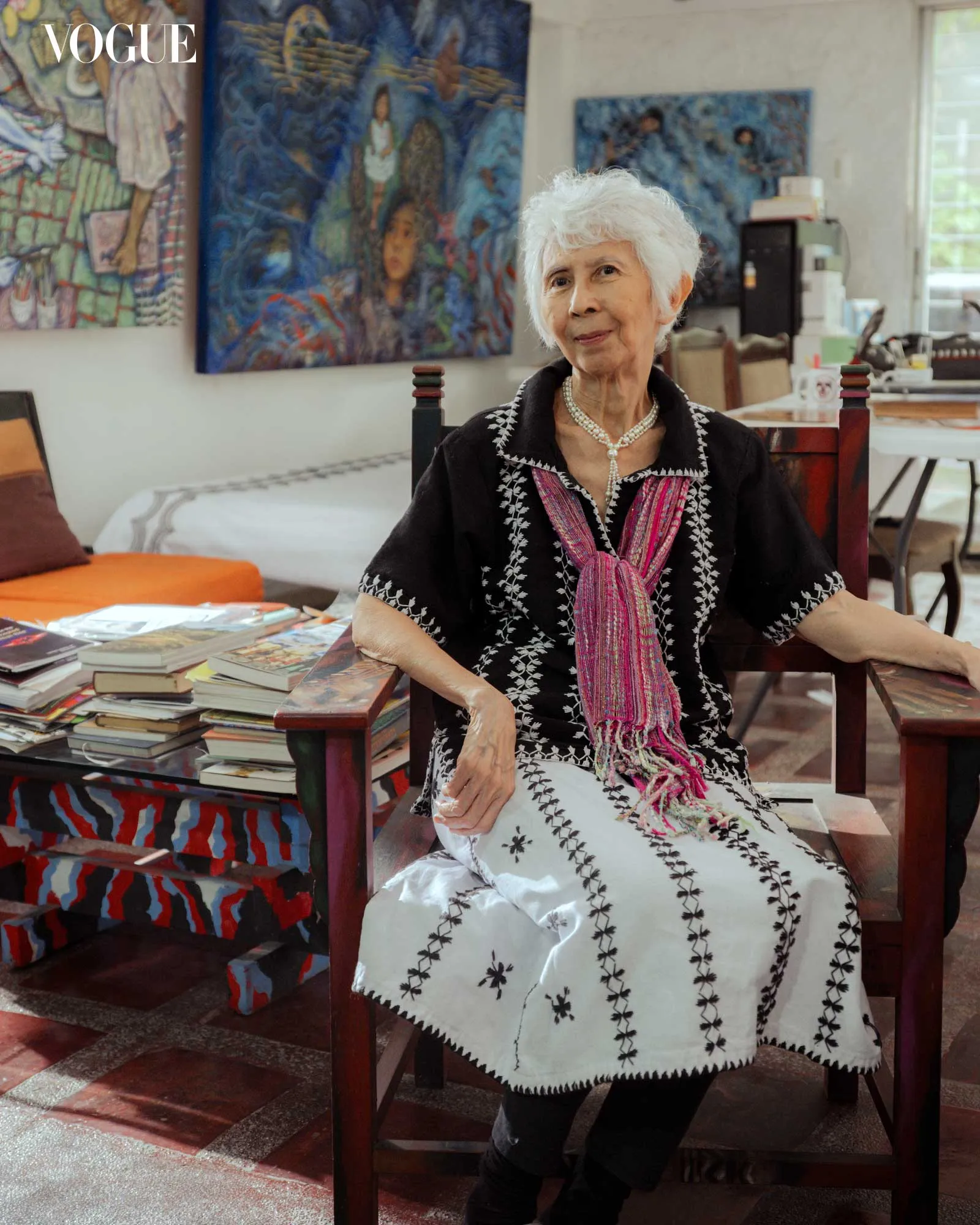 Portrait of an elderly woman with short white hair, wearing a black embroidered dress and pink scarf, seated in a room filled with colorful paintings and books by a table.