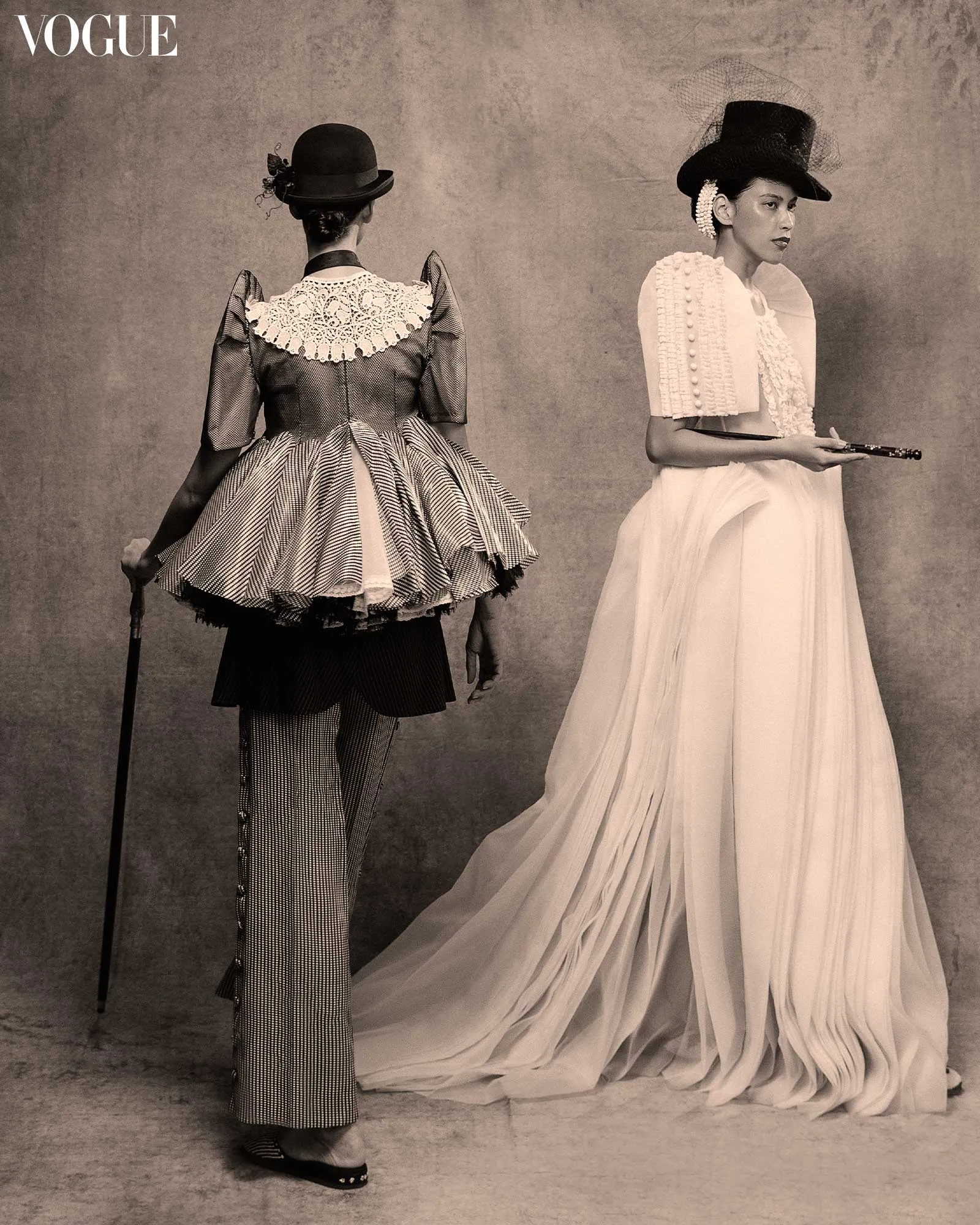 Two women in vintage gowns pose side by side: one with lace collar facing away, the other in a white gown with a black hat facing right, on a textured backdrop for Vogue.