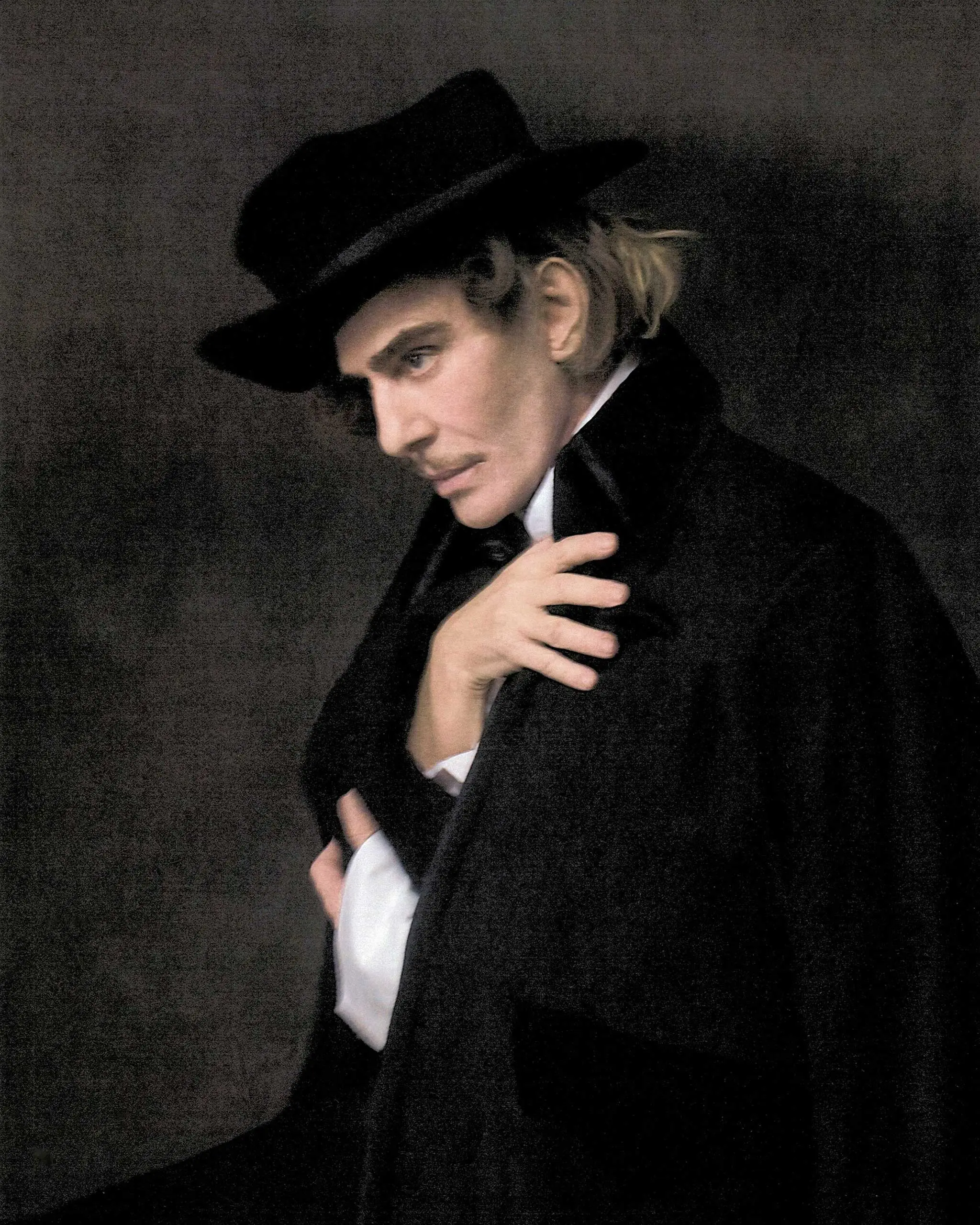 Portrait of a person wearing a black wide-brim hat and dark coat, looking down against a dark background.