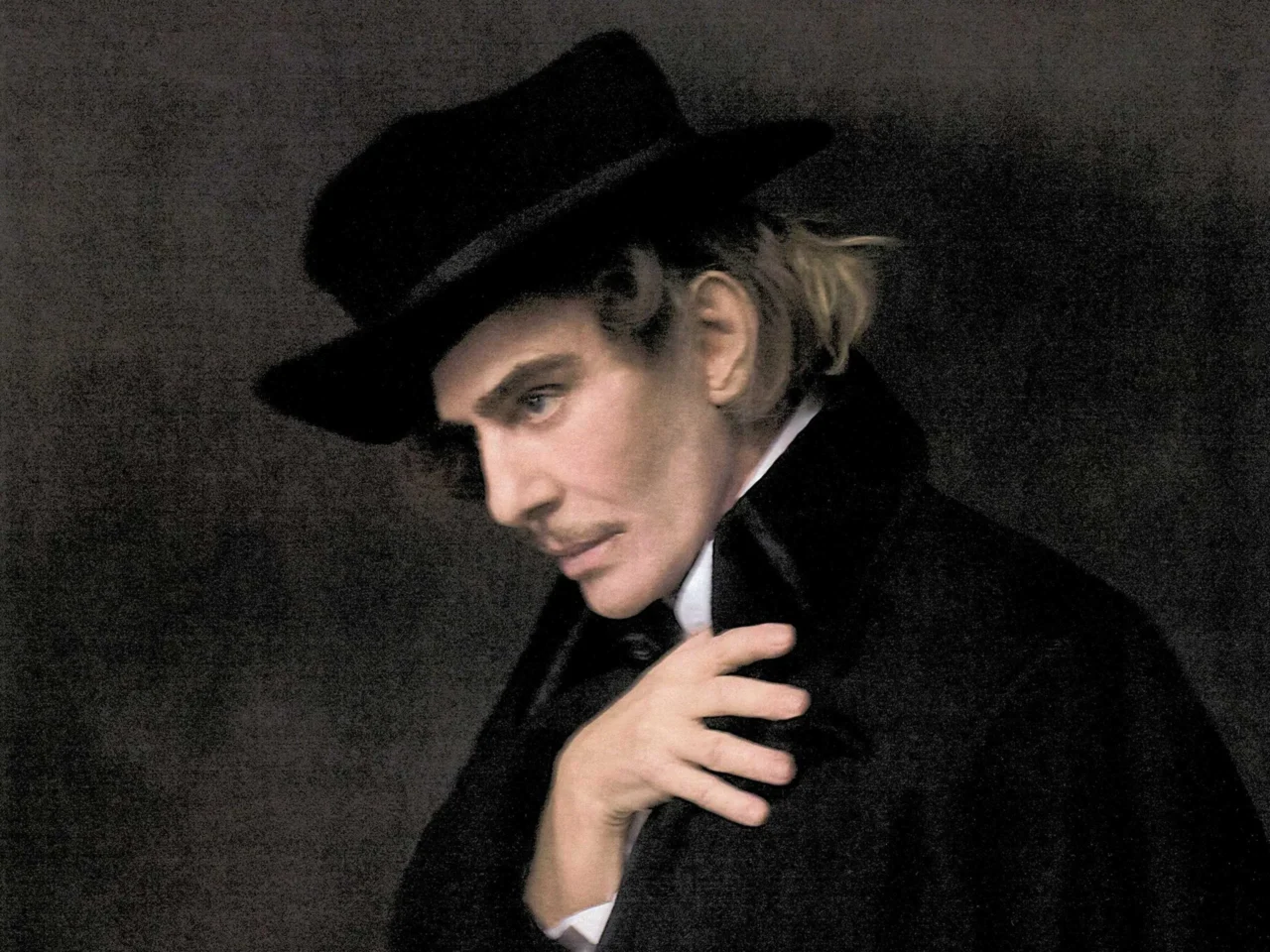 Person wearing a black fedora and tuxedo, looking downward against a dark backdrop.