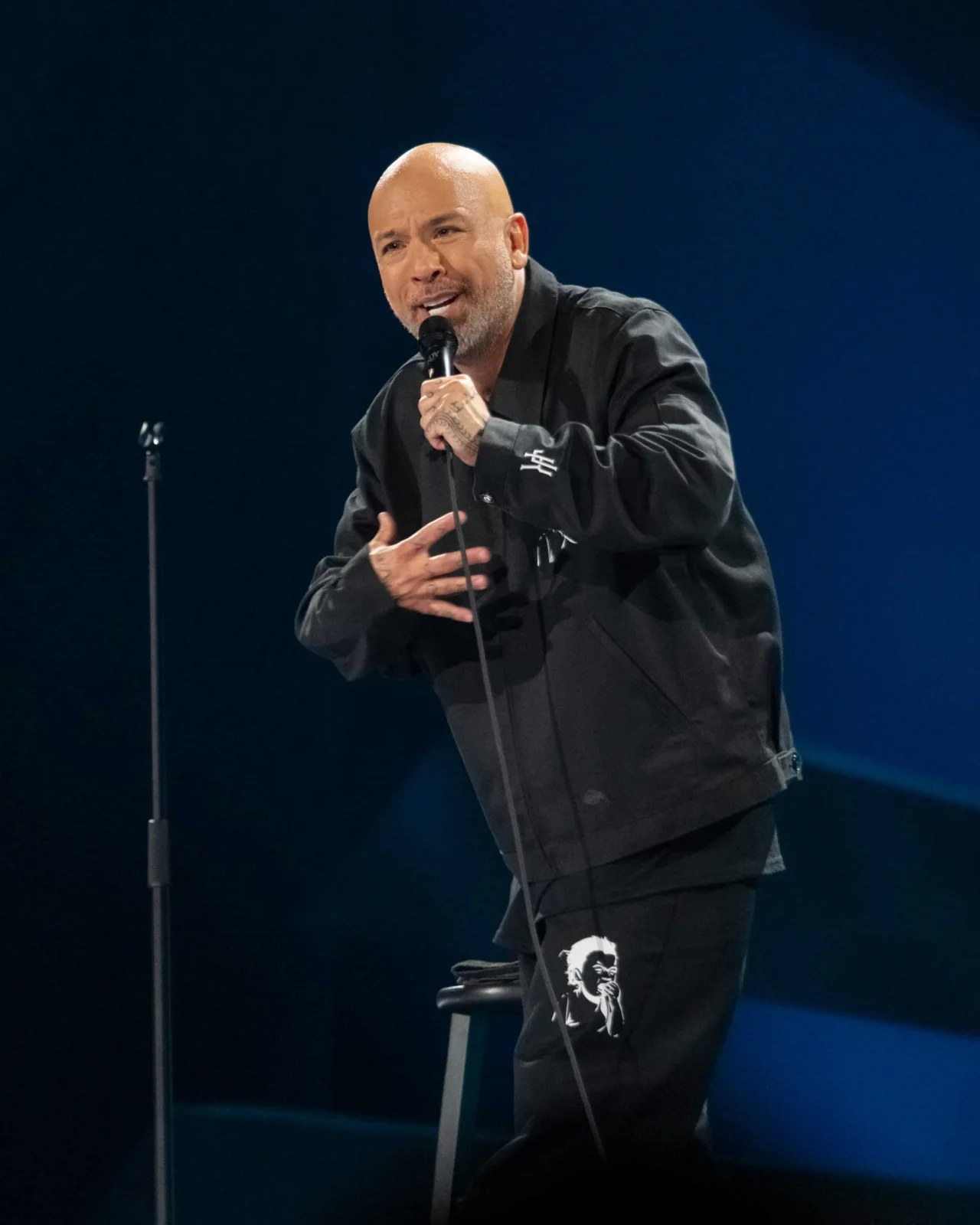 Comedian performing a stand-up set on stage, speaking into a handheld microphone.