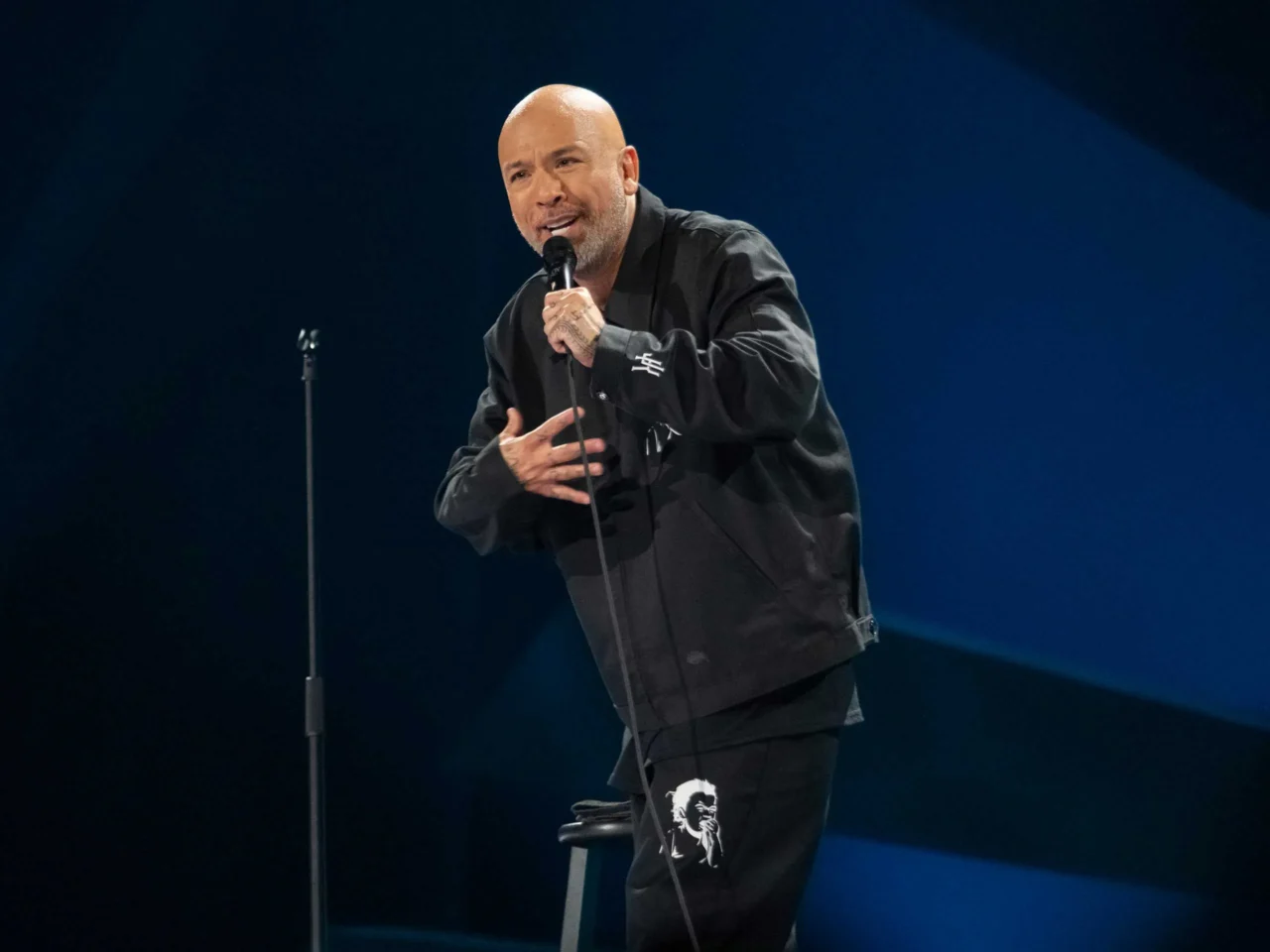 Comedian performing a stand-up set on stage, speaking into a handheld microphone.