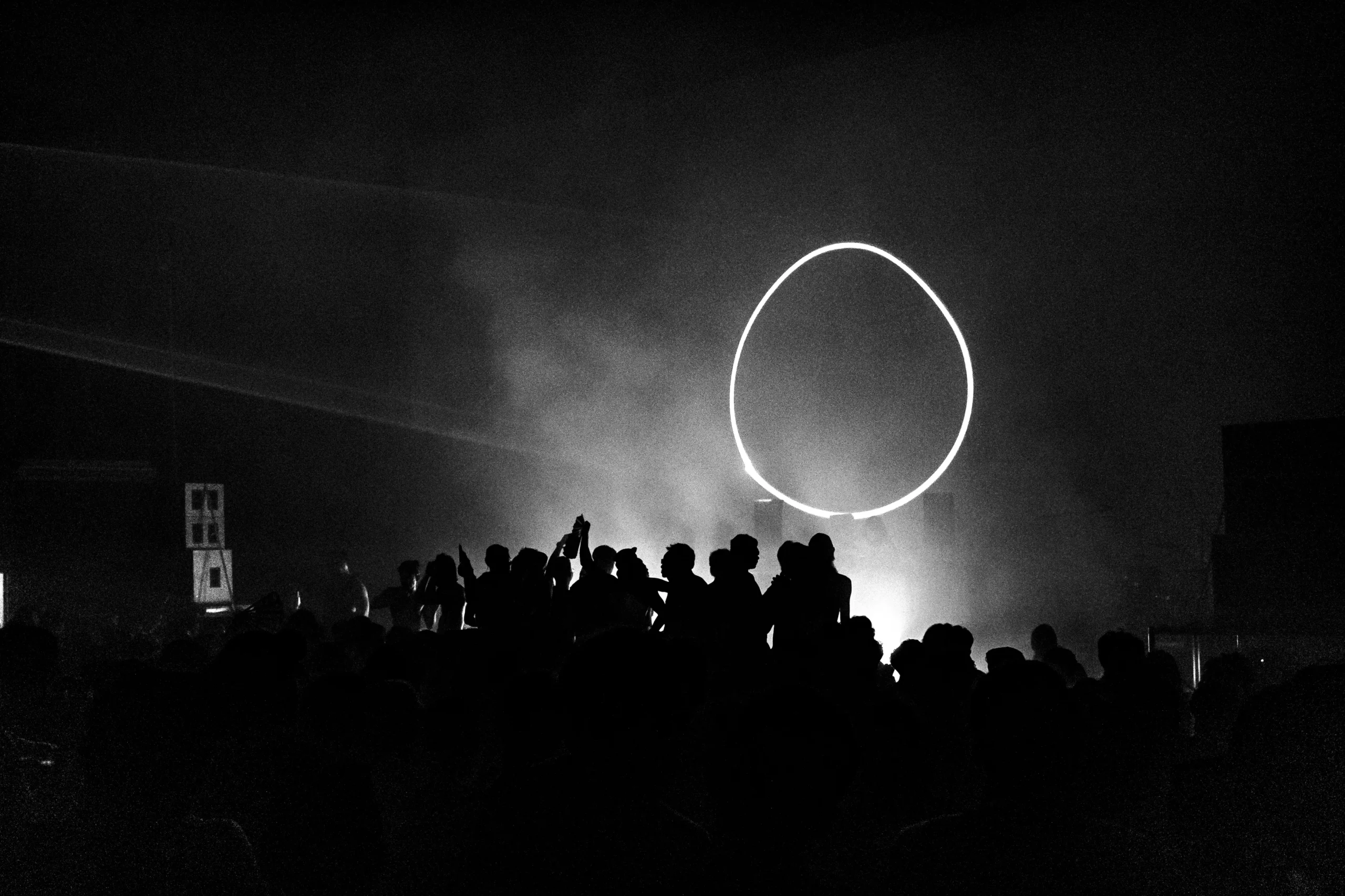 Silhouetted crowd at a dark concert with a bright circular light ring in the air above them.