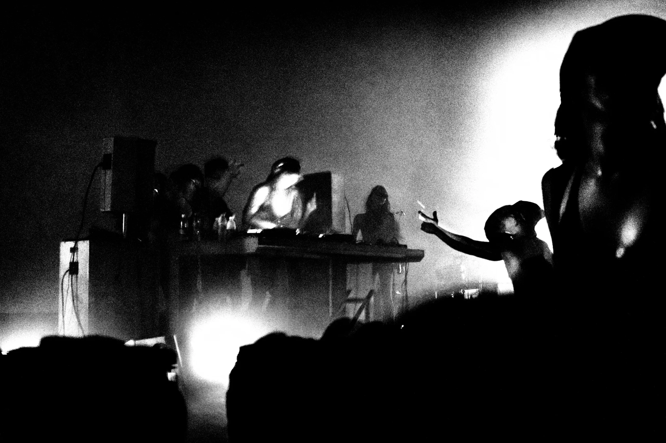 DJ performing at a booth on a dark stage, with silhouetted crowd in front and hazy lights behind.