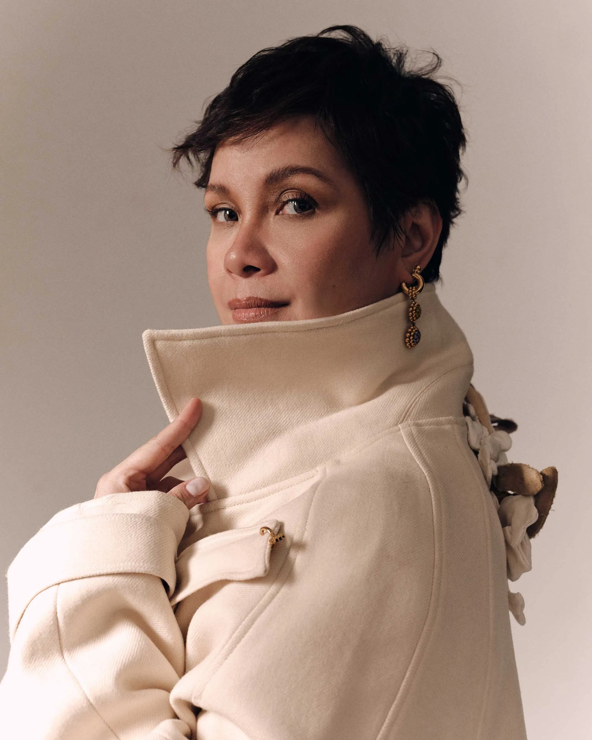 Portrait of a person with short dark hair wearing a beige high-collar coat and gold earrings, looking to the side.