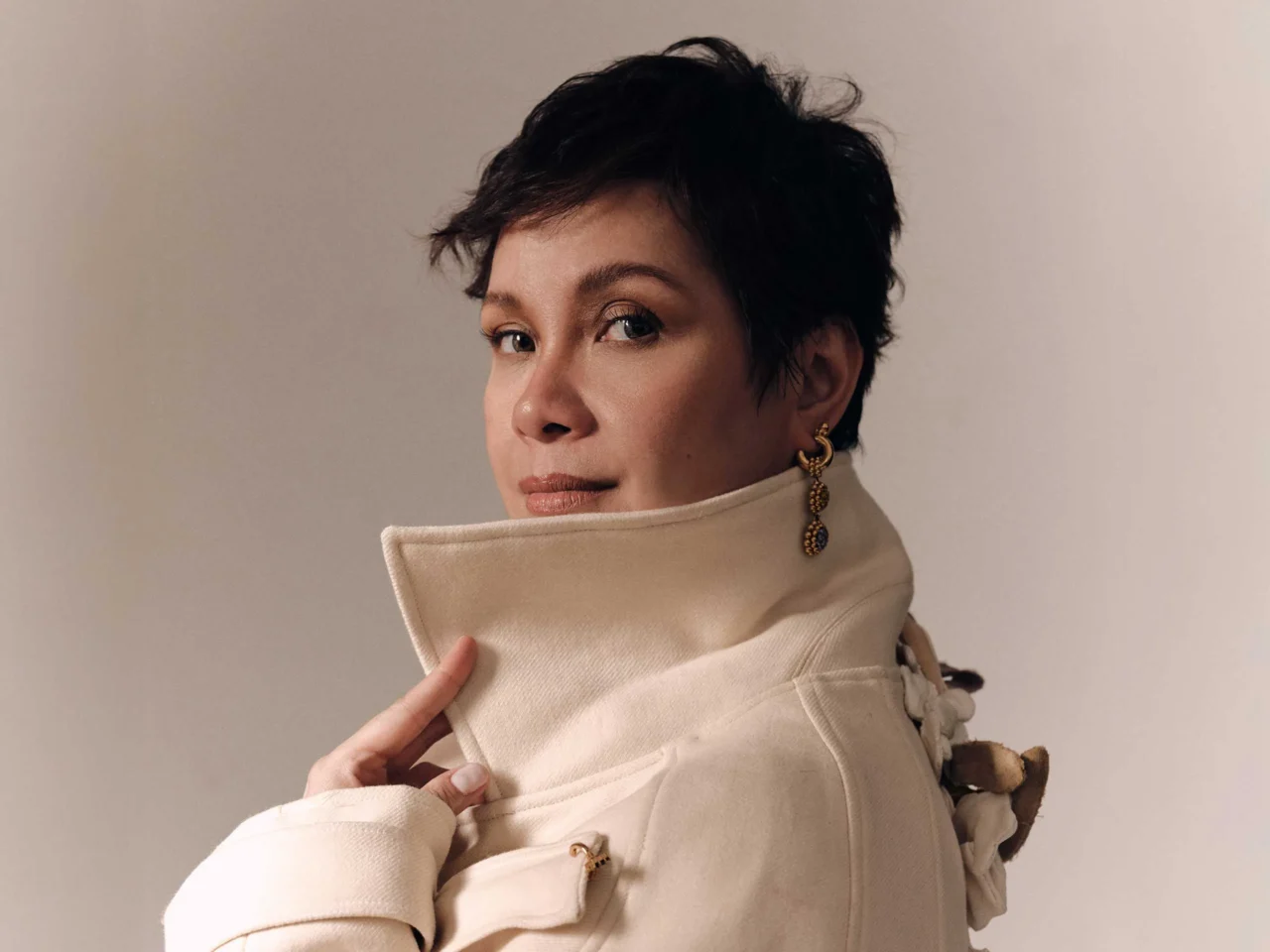 Portrait of a person with short dark hair wearing a beige high-collar coat and gold earrings, looking at the camera.