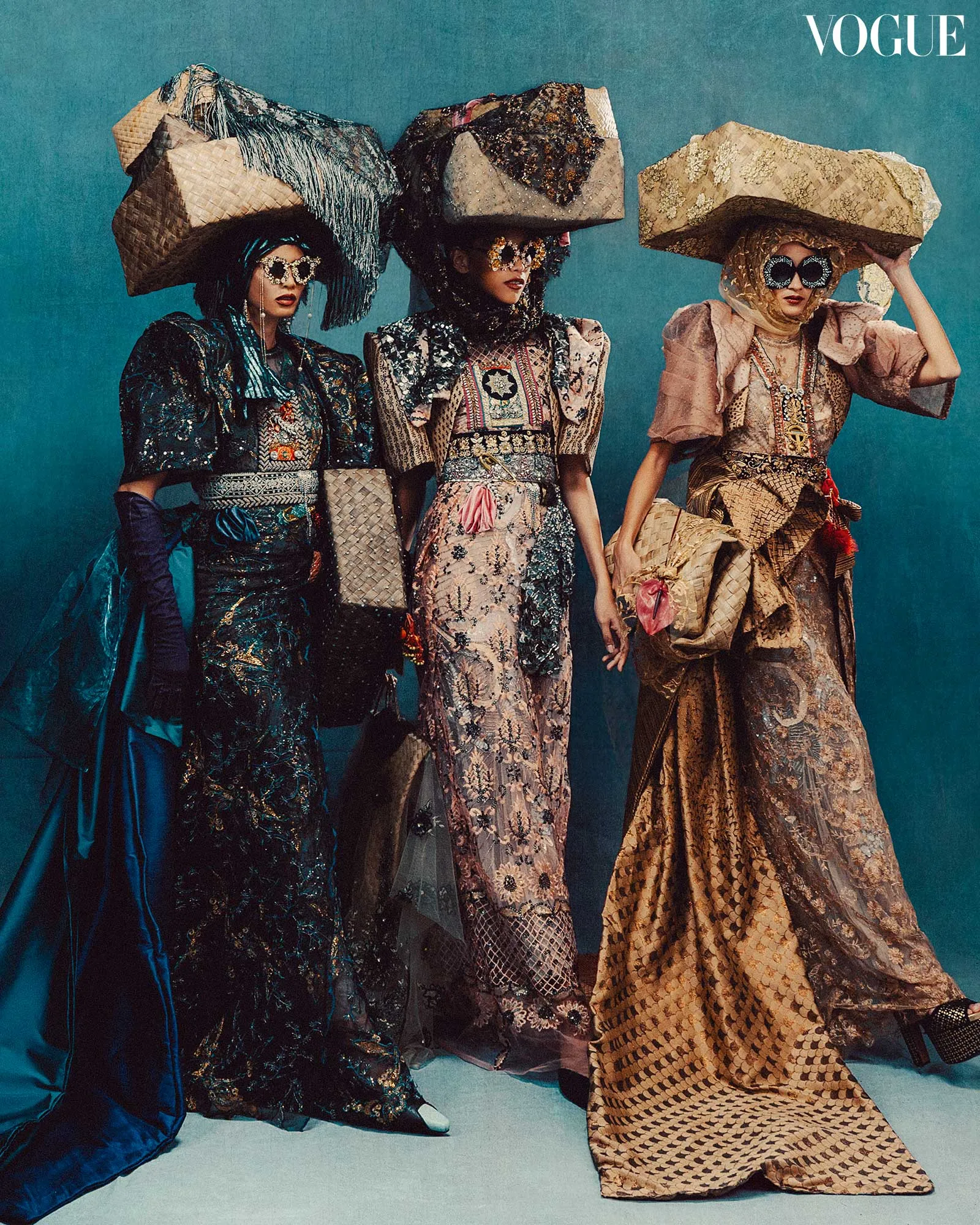 Three models in ornate, jewel‑embellished gowns wear oversized woven hats and sunglasses against a teal backdrop for a Vogue editorial.
