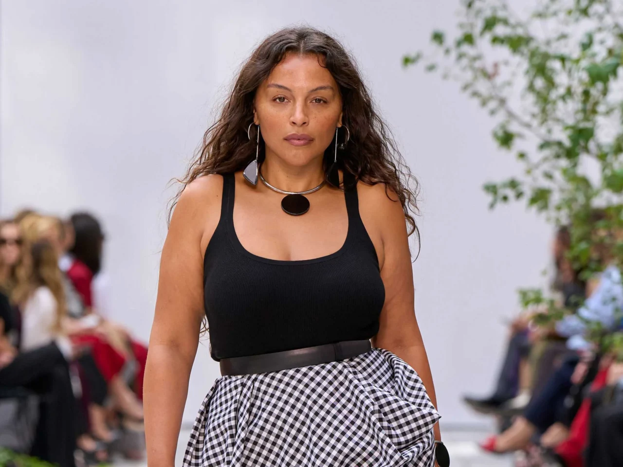 Model walking a runway in a black sleeveless V-neck dress with a silver choker and bold red lipstick, blurred spectators in the background.