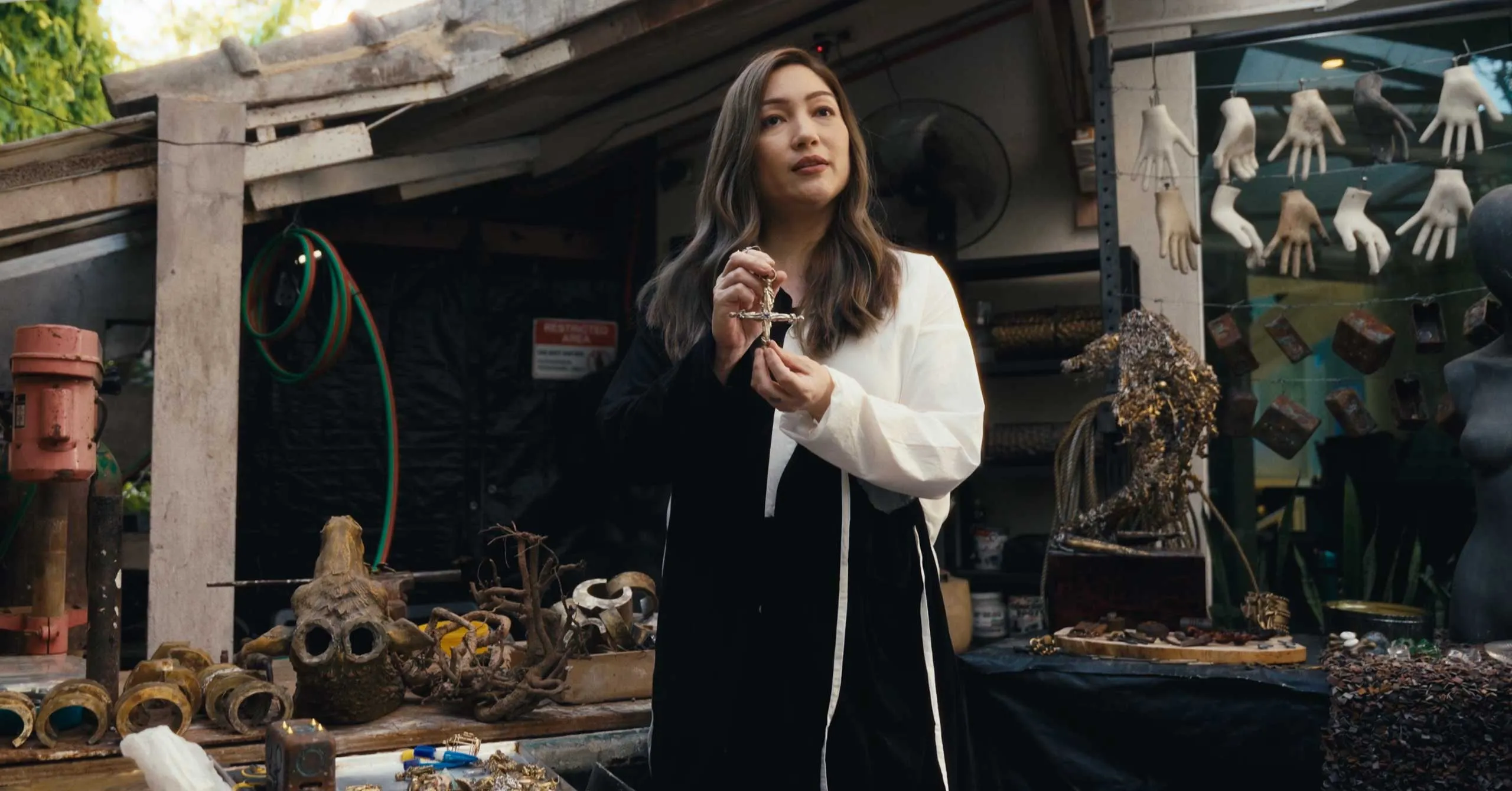 Woman in a black and white coat holds a cross pendant at a market stall displaying skulls, bones, and trinkets behind her.