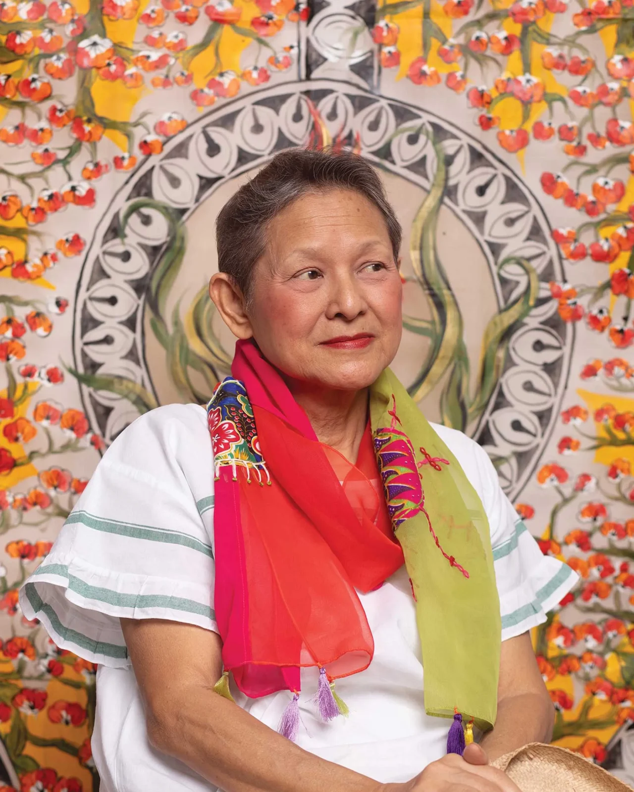 Portrait of a smiling woman wearing a bright, multicolored scarf, looking to the right against a floral mural backdrop.