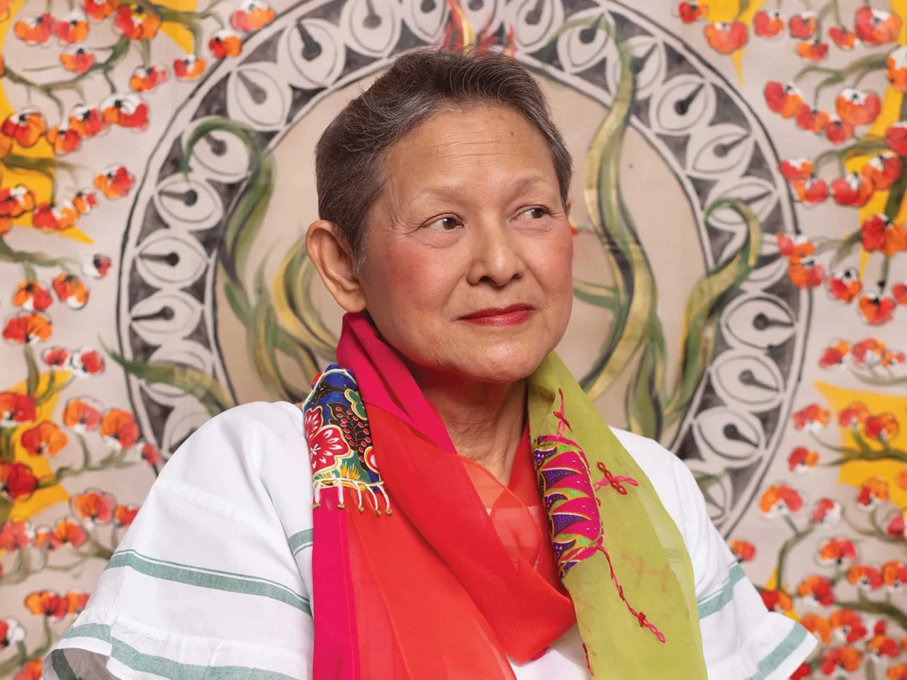 Portrait of a smiling woman wearing a bright, multicolored scarf, looking to the right against a floral mural backdrop.