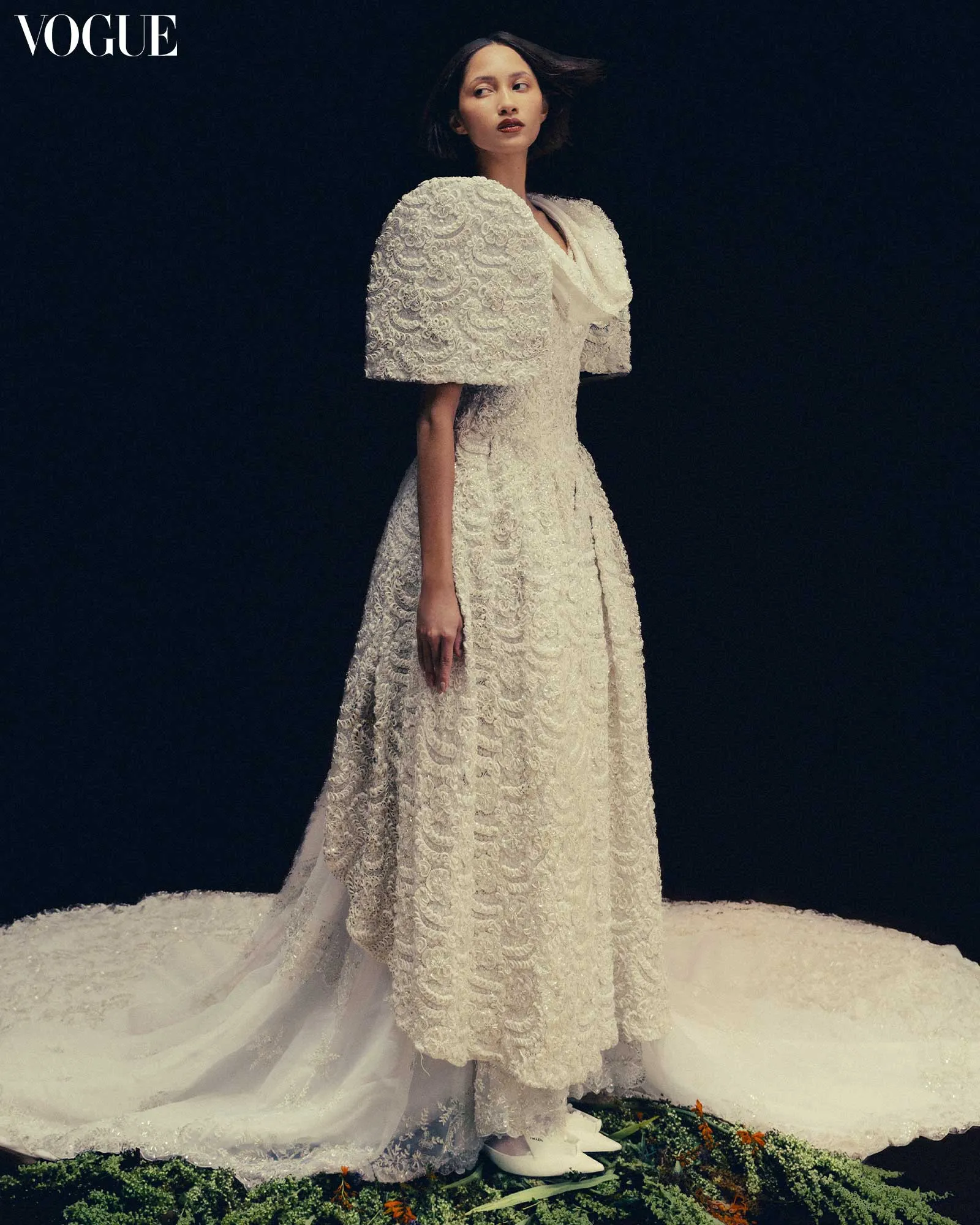 Model wearing an intricate white lace wedding gown with dramatically sculpted sleeves, standing against a dark backdrop on a snowy runway.