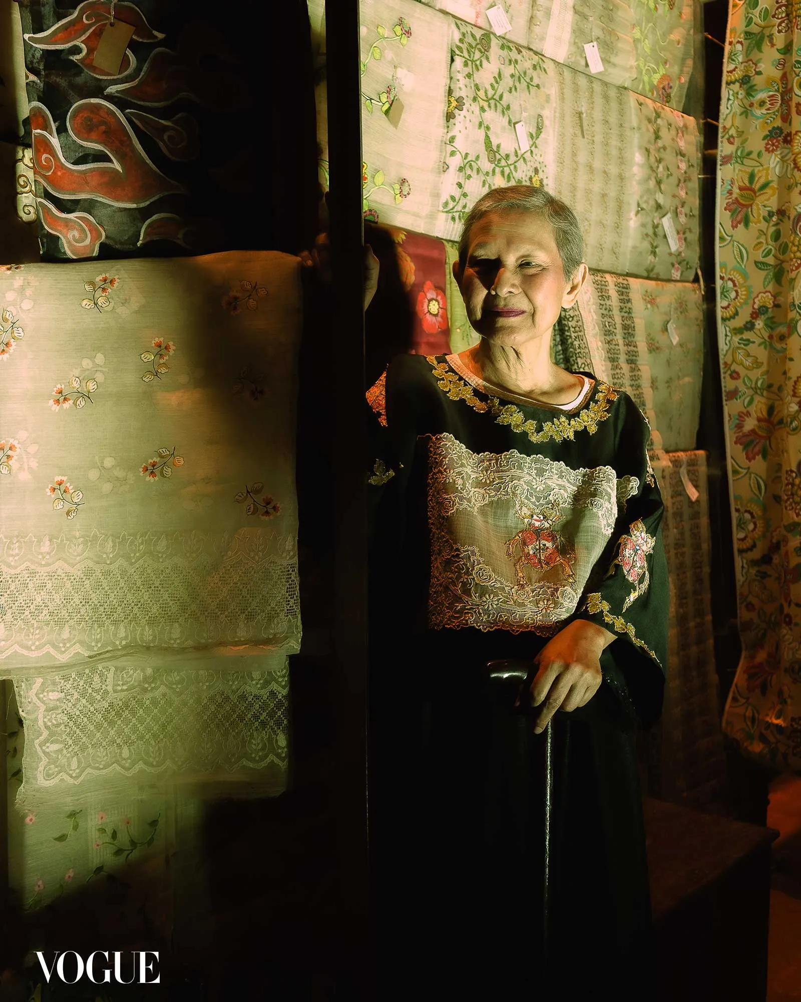 Older woman in a black and gold embroidered dress sits among stacked patterned fabrics in a dim textile shop