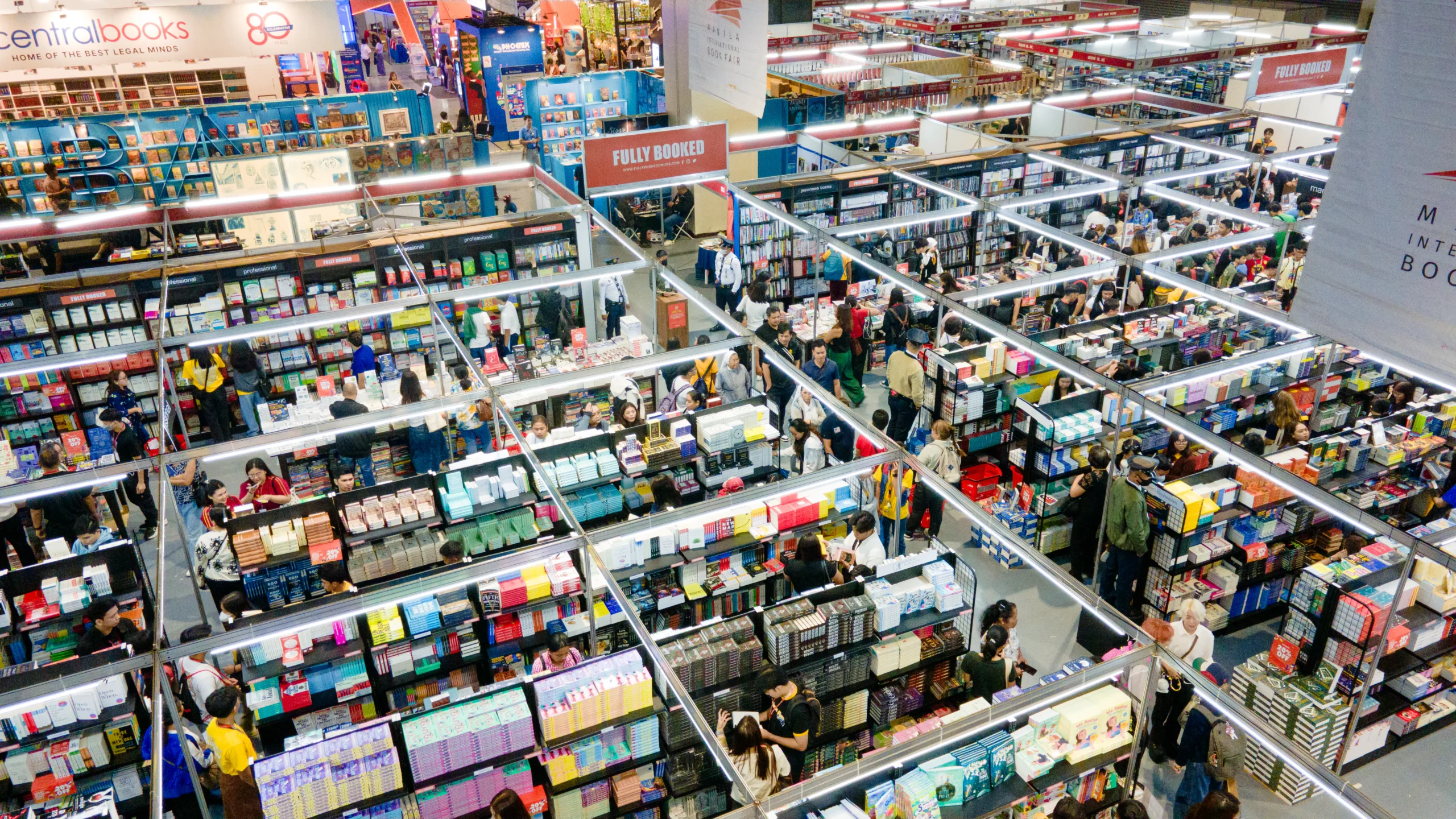 Overhead view of a crowded book fair with tall shelves, bright signage, and many shoppers browsing books on display shelves.