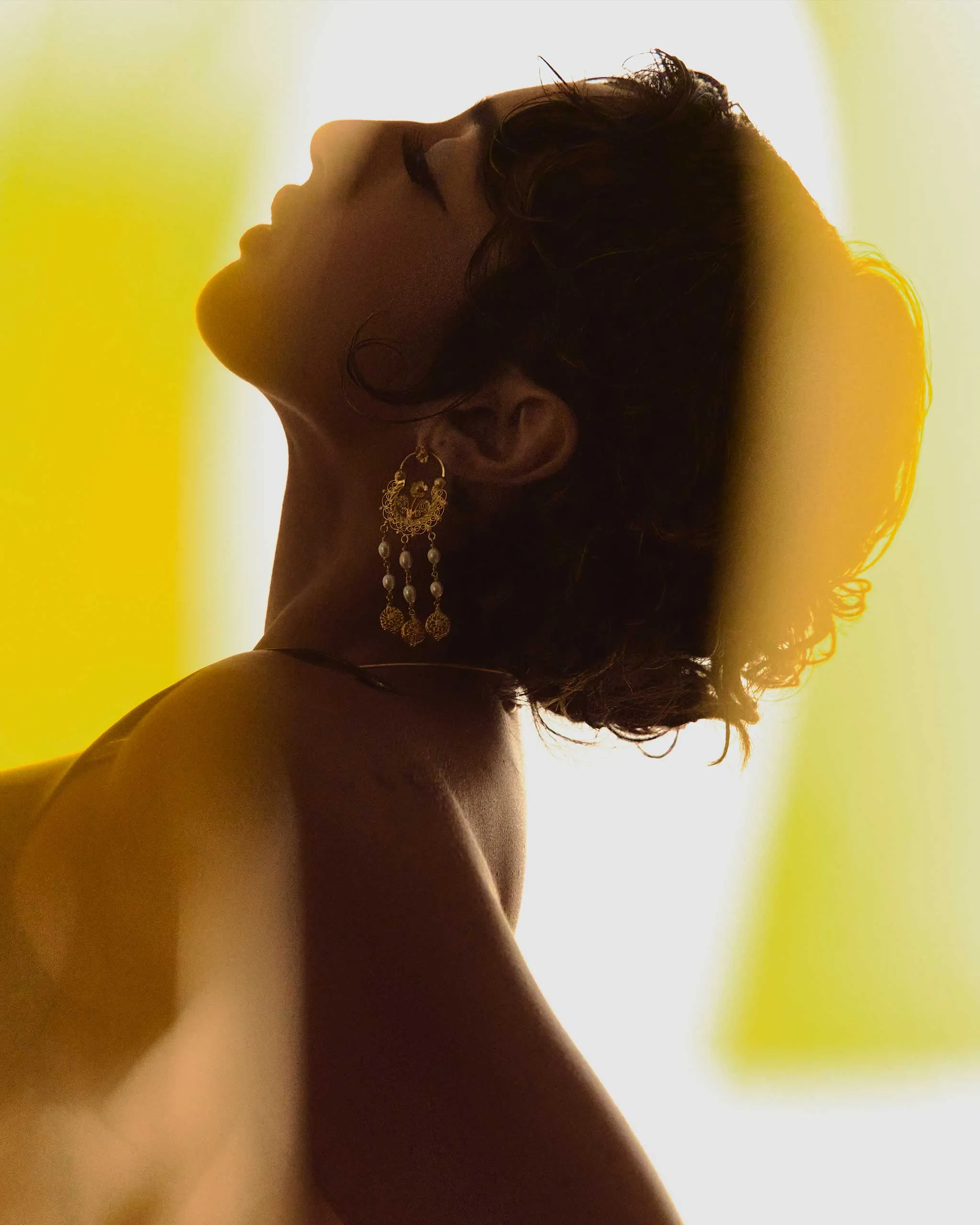 Profile of a woman with head tilted back, wearing ornate dangling earrings, against a bright yellow backlight.