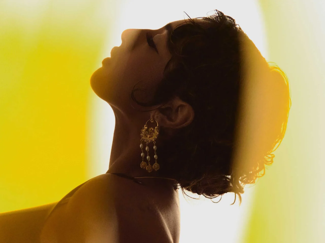 Profile of a woman looking upward, wearing ornate gold chandelier earrings against a bright yellow background.