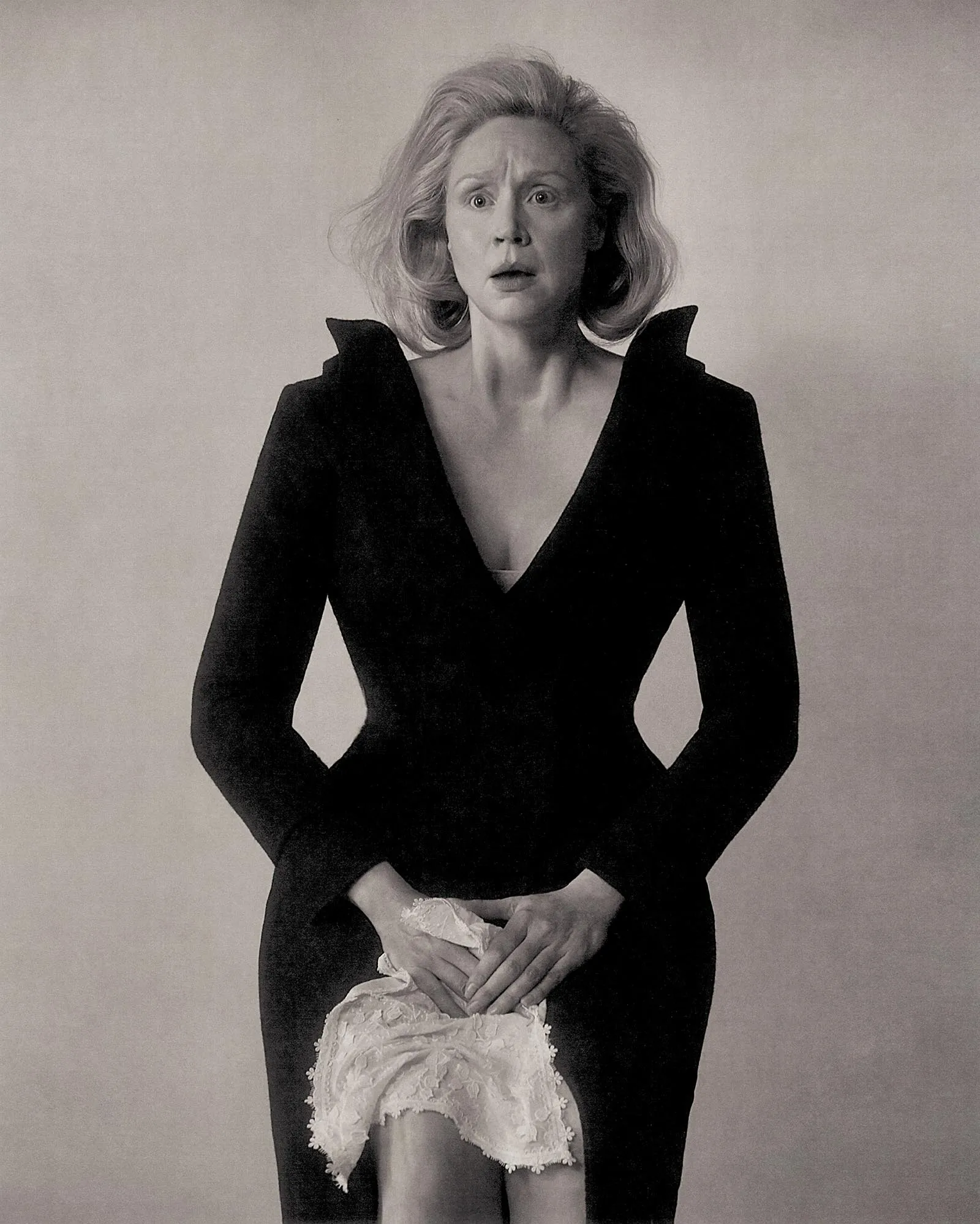 Black-and-white photo of a surprised woman in a deep V-neck black dress, holding a lace cloth in front of her.