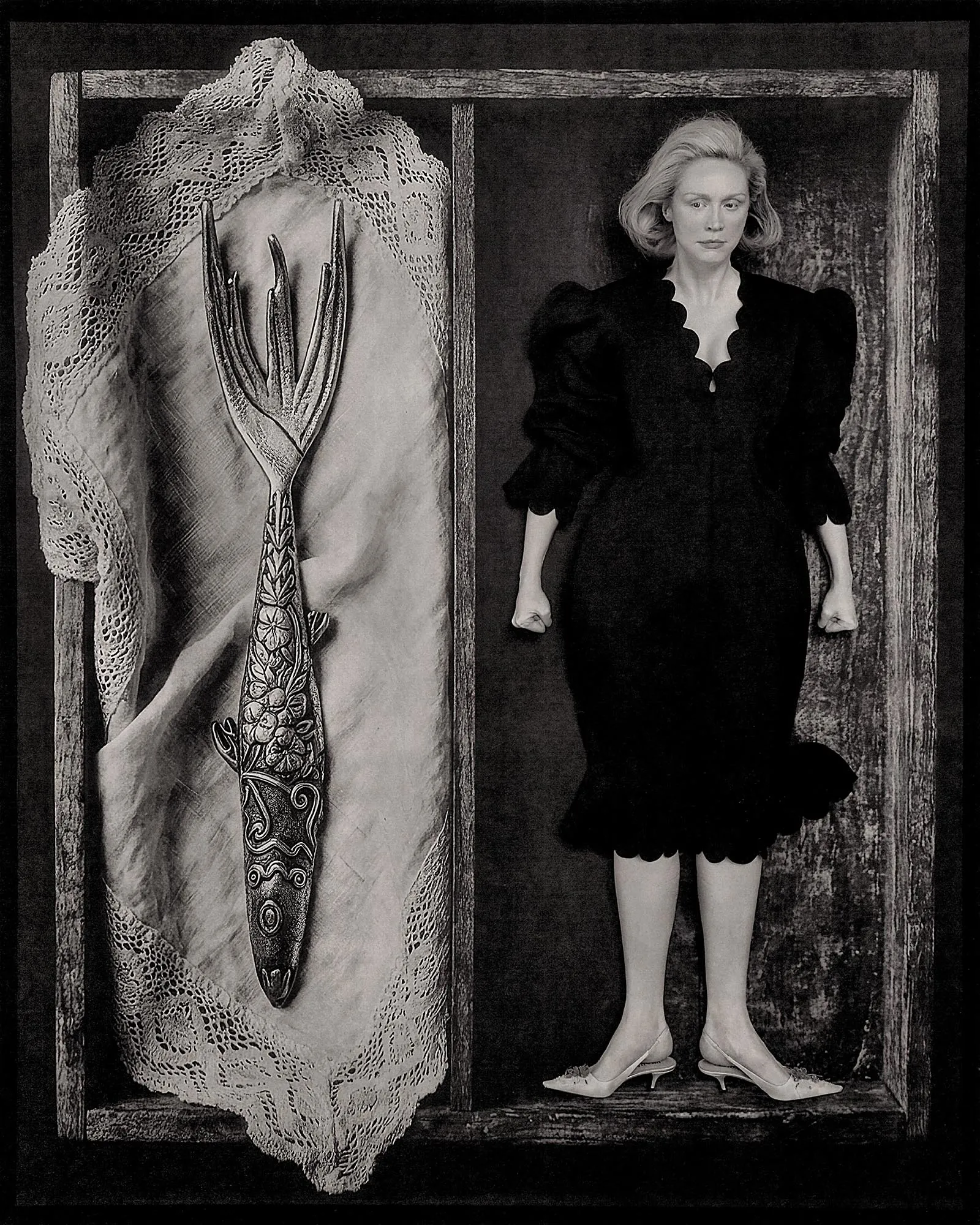 Black-and-white photo of a woman in a vintage black dress standing inside a wooden frame, with a carved, dagger-like object wrapped in lace to her left.