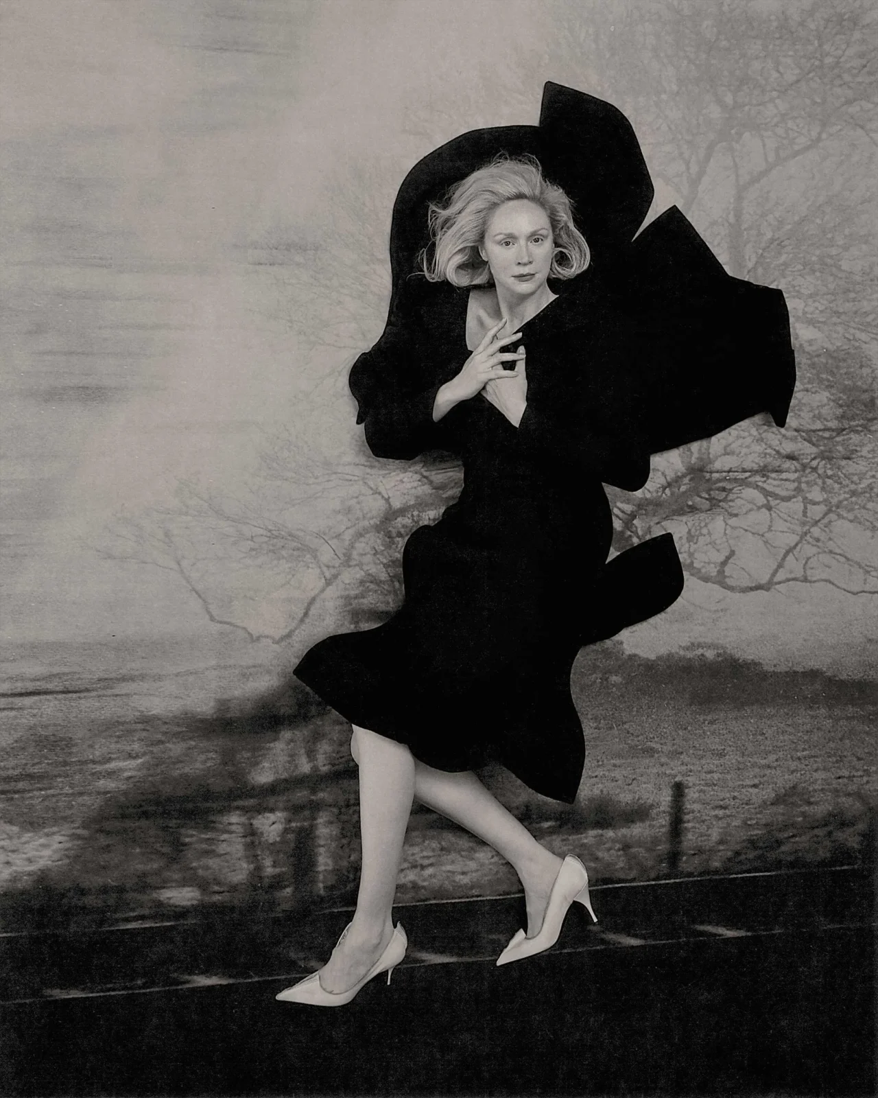 Black-and-white portrait of a woman in a dramatic oversized black cape, her hands clasped near her chest, posing against a textured backdrop.