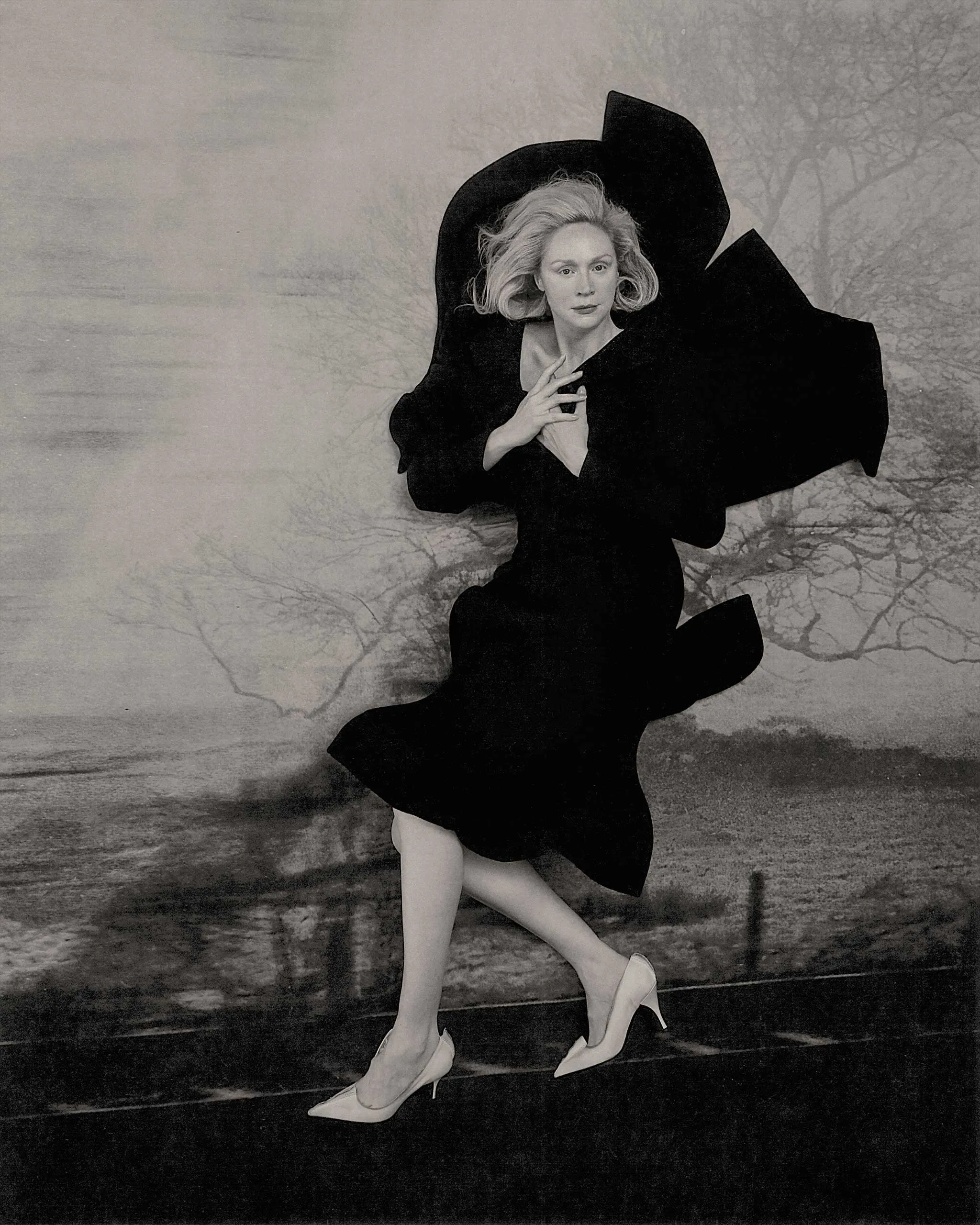 Black-and-white photo of a woman in a flowing black dress and high heels, posing as if dancing against a painted backdrop.