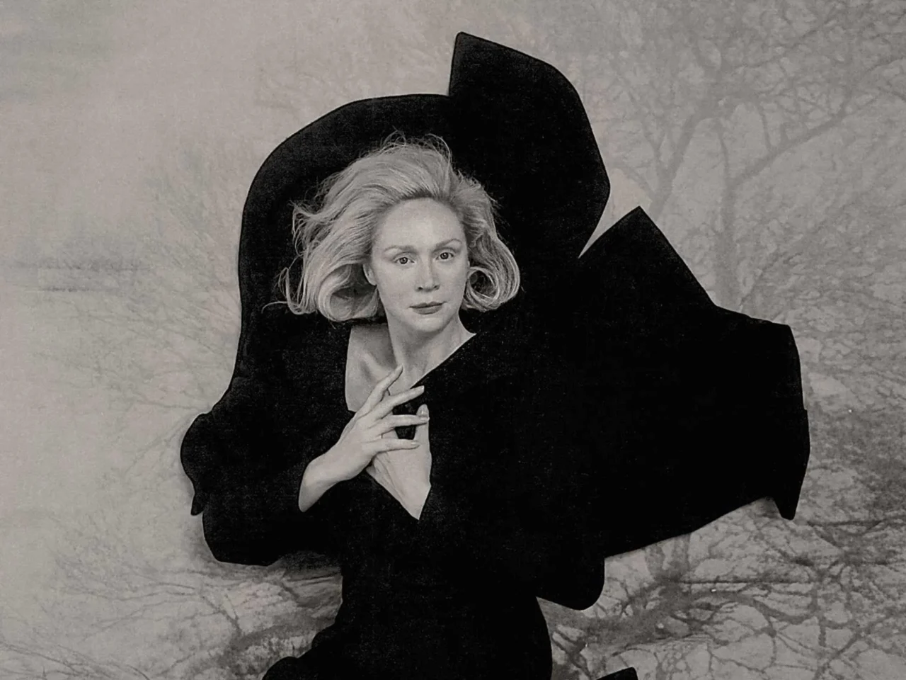 Black-and-white portrait of a woman in a dramatic oversized black cape, her hands clasped near her chest, posing against a textured backdrop.