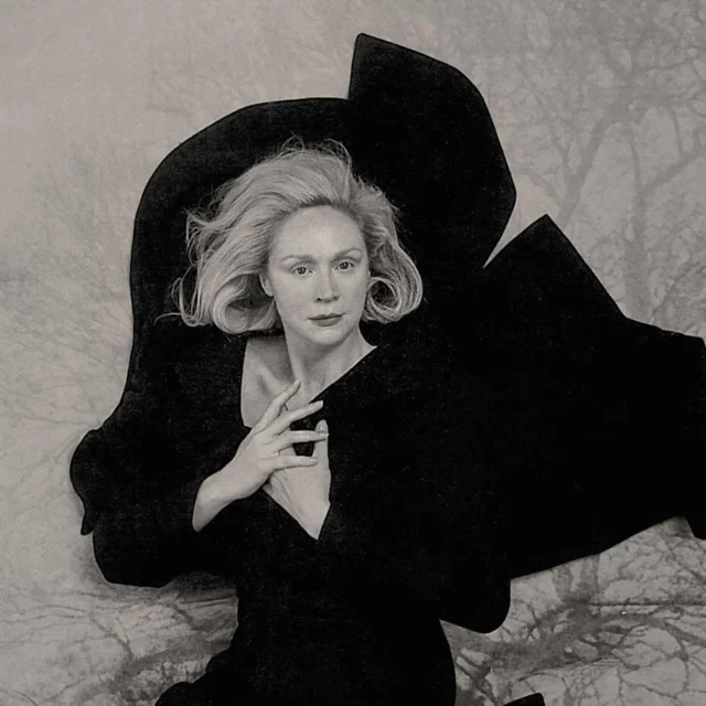 Black-and-white portrait of a woman in a large black hat and a dark dress, posing with hands near her chest against a marbled backdrop.