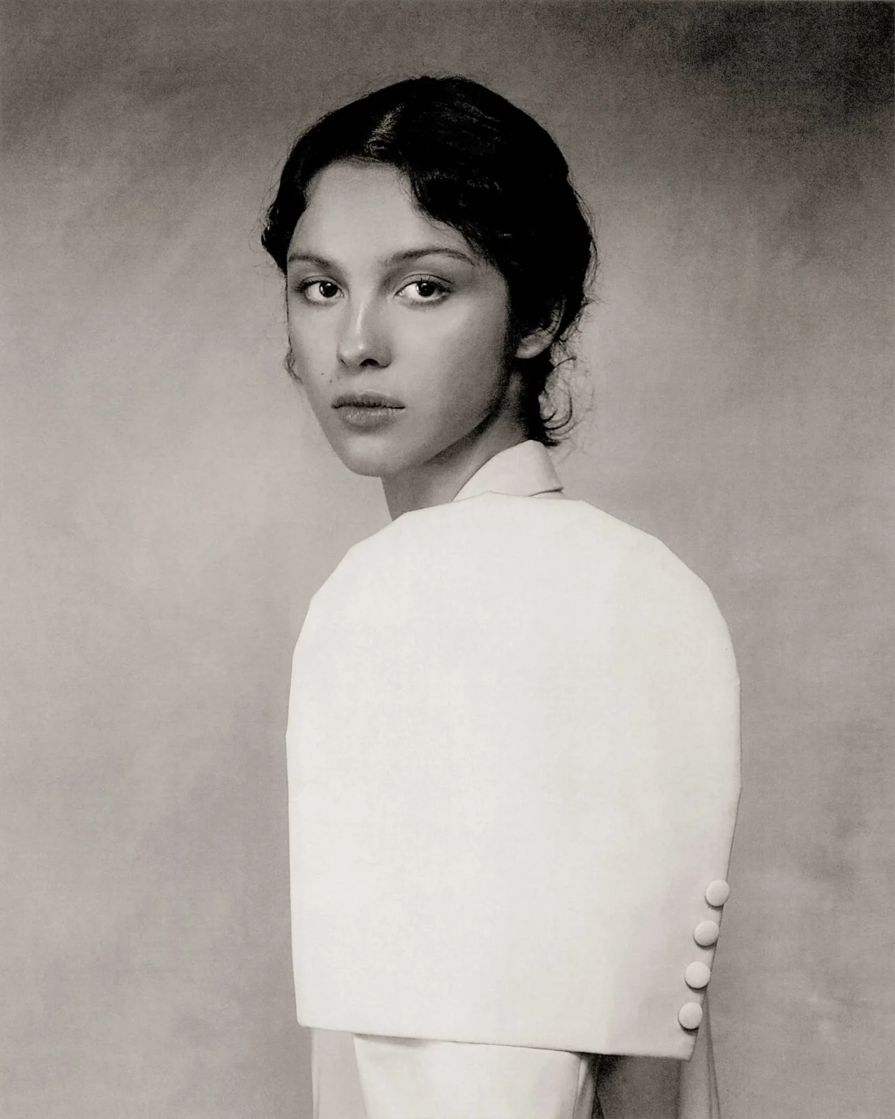 Black-and-white portrait of a woman looking over her shoulder, soft expression, cropped shoulders visible behind.
