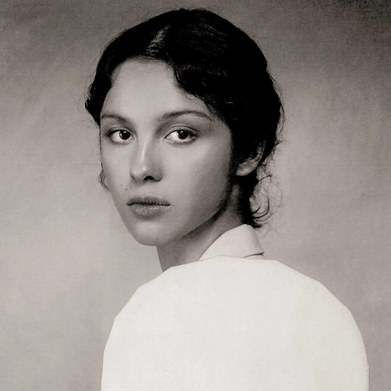 Black-and-white portrait of a woman looking over her shoulder, soft expression, cropped shoulders visible behind.