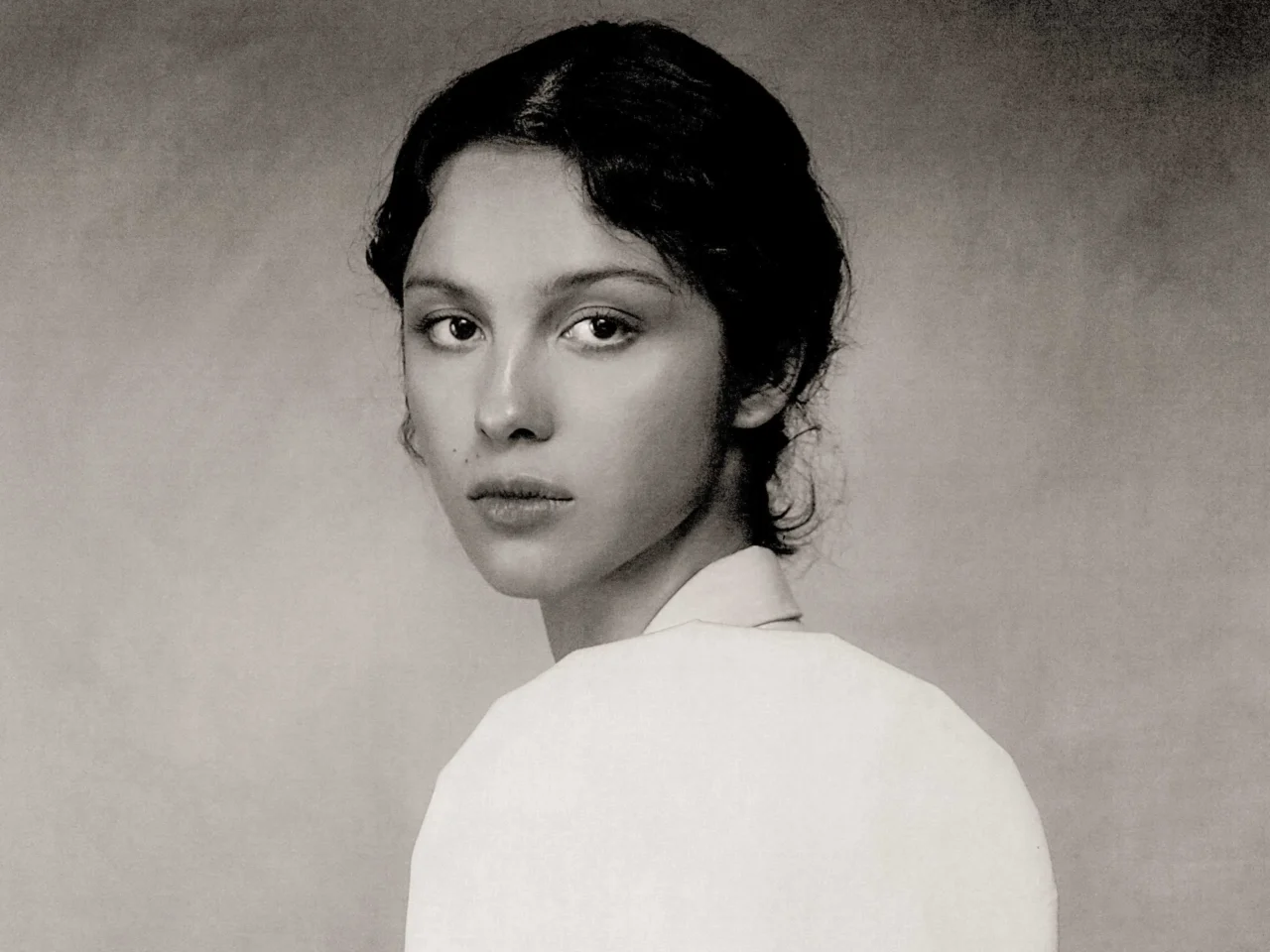 Black-and-white portrait of a woman looking over her shoulder, soft expression, cropped shoulders visible behind.