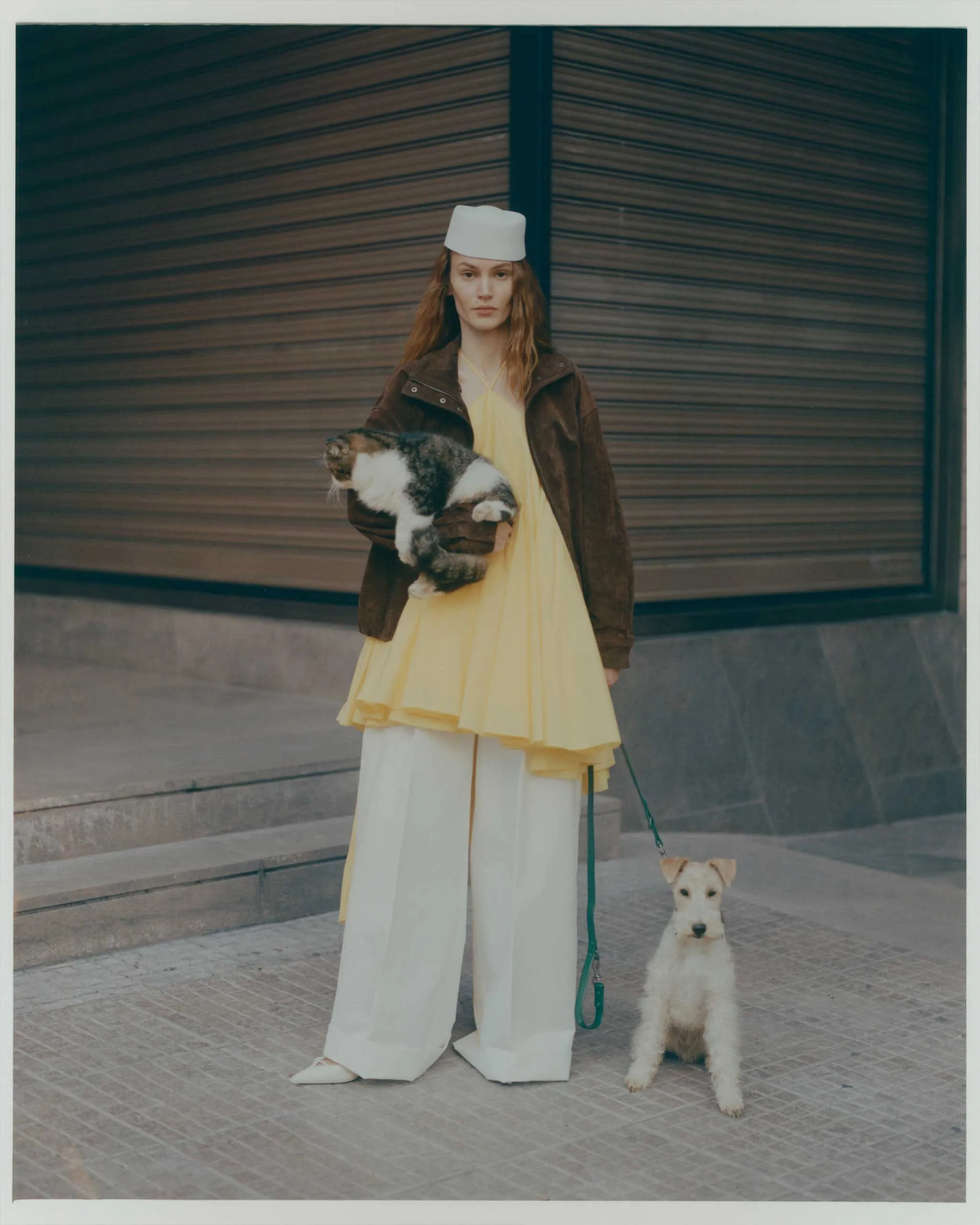 Woman in a pale yellow dress and brown coat with a white hat stands on a sidewalk, holding a small fluffy dog; another white dog on a leash sits nearby in front of a metal shutter.
