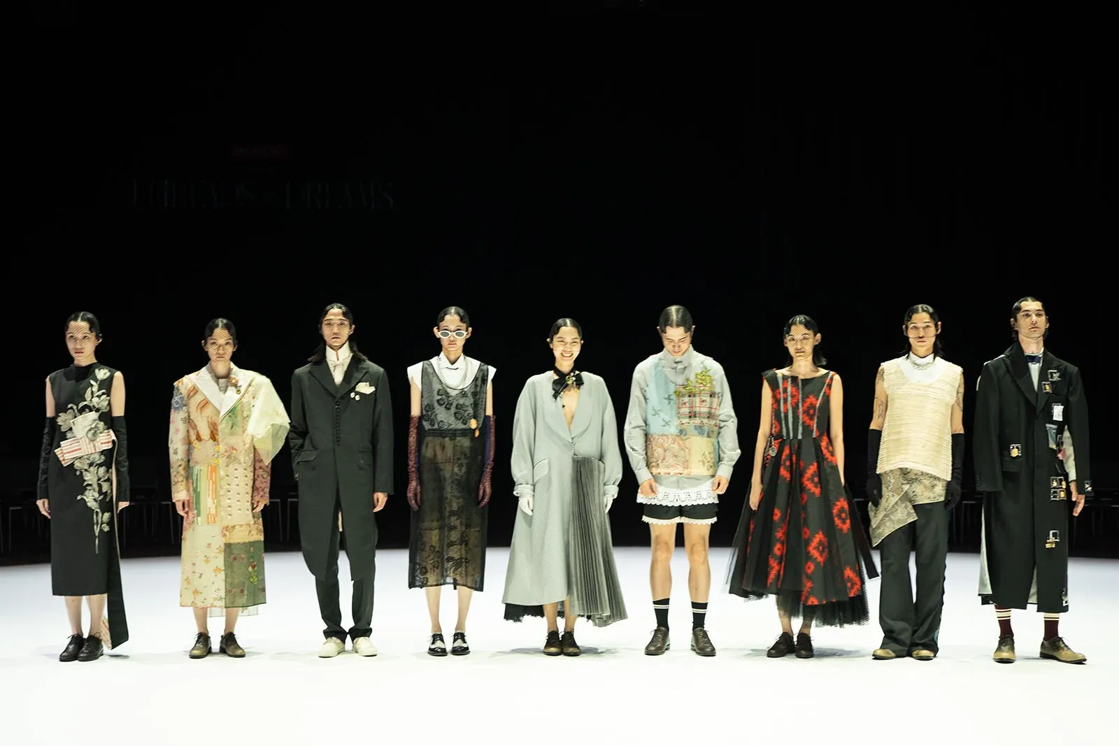 Nine models stand in a line on a white runway, wearing varied avant-garde outfits against a dark backdrop at a fashion show.