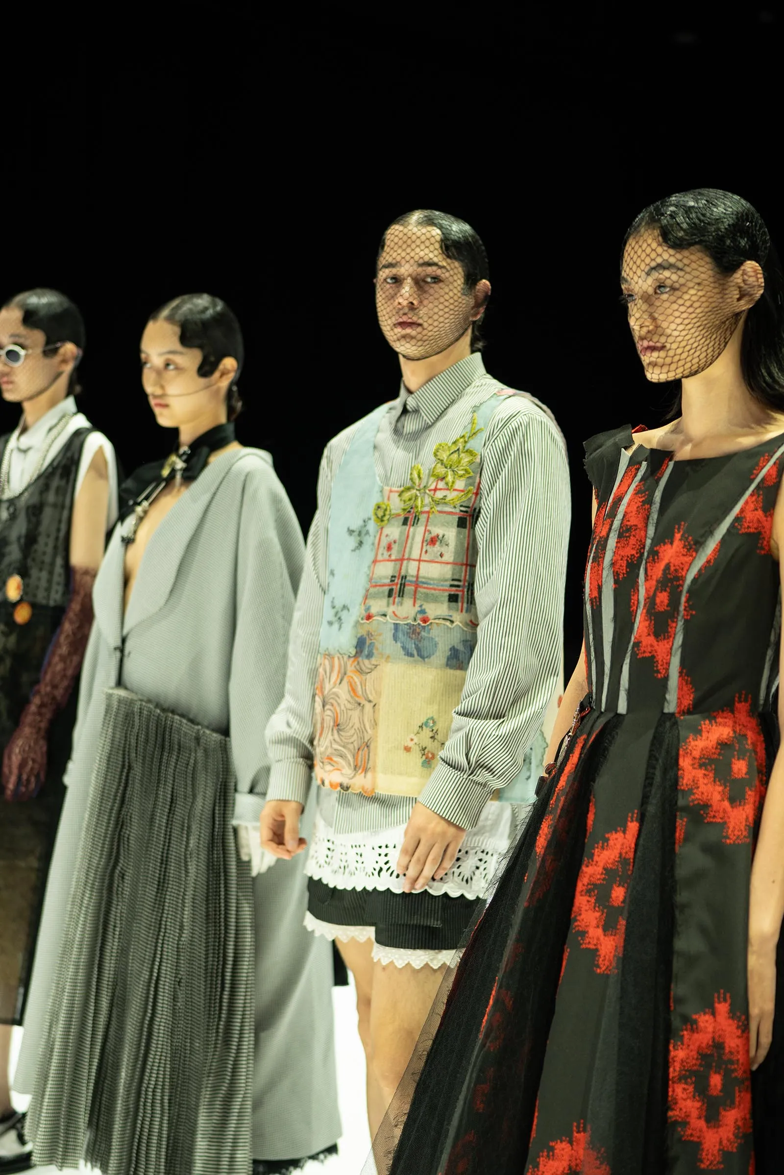 Models line up on a runway wearing eclectic outfits, many with mesh veils over their faces and bold patterns in muted tones and red accents.