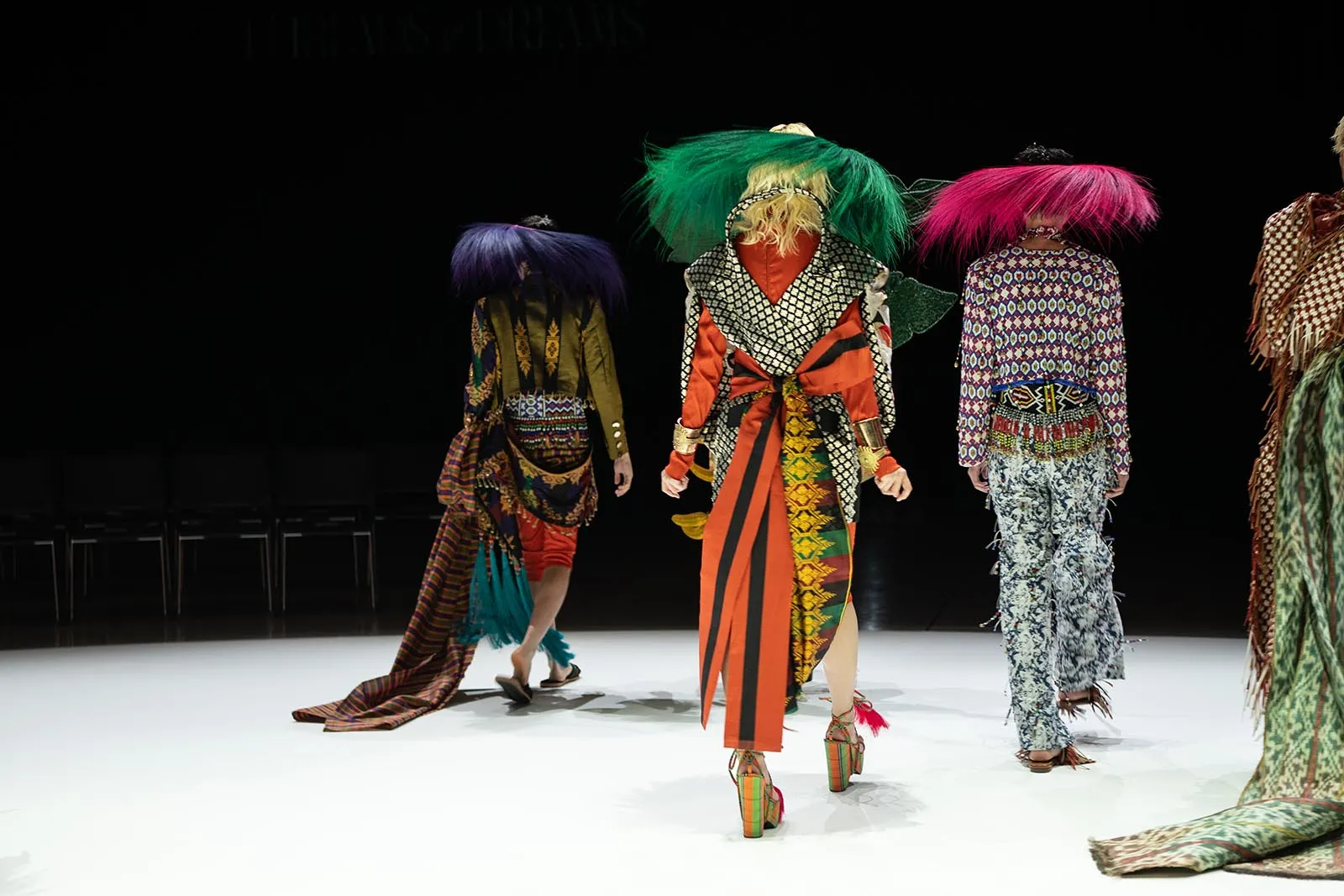 Models walk away from the camera on a white runway, wearing bold patterned outfits with large colorful feathered hats in green, purple, and pink.