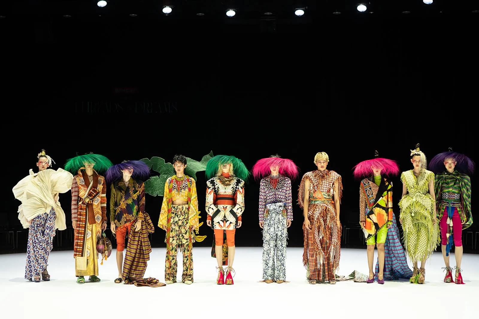 A row of models on a runway wearing bright, avant-garde outfits with bold patterns and oversized accessories under stage lights.