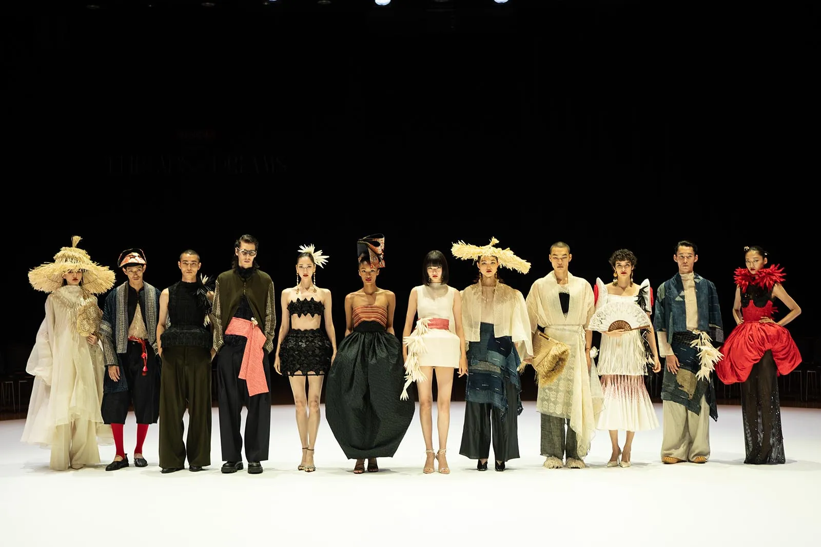 A line of fashion models in avant-garde outfits stands on a white runway against a dark backdrop, showcasing dramatic hats, sculptural dresses, and bold styling.