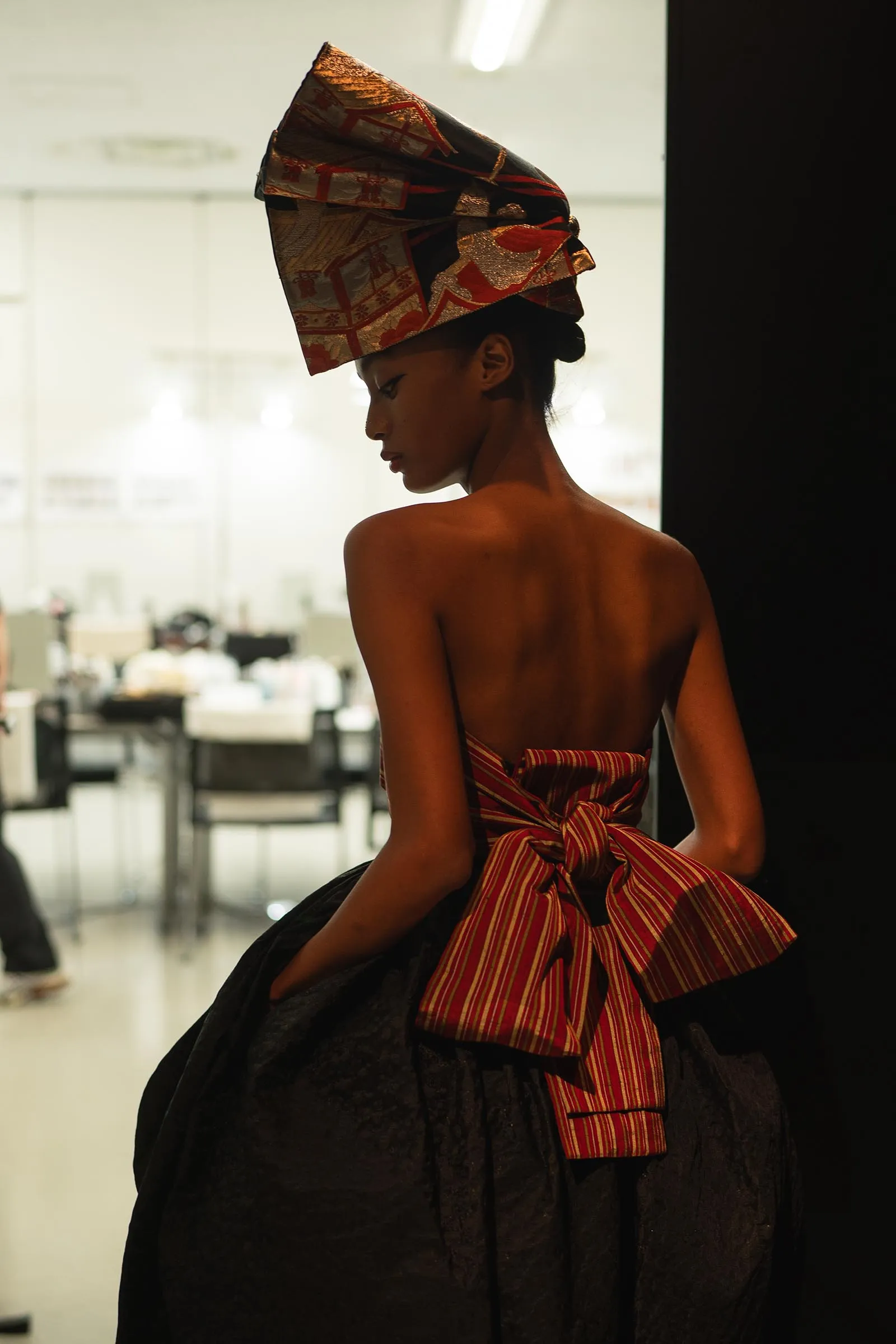 Profile of a woman in a large, ornate red-and-gold headwrap and a black dress with a red-striped sash tied at the back, facing left.