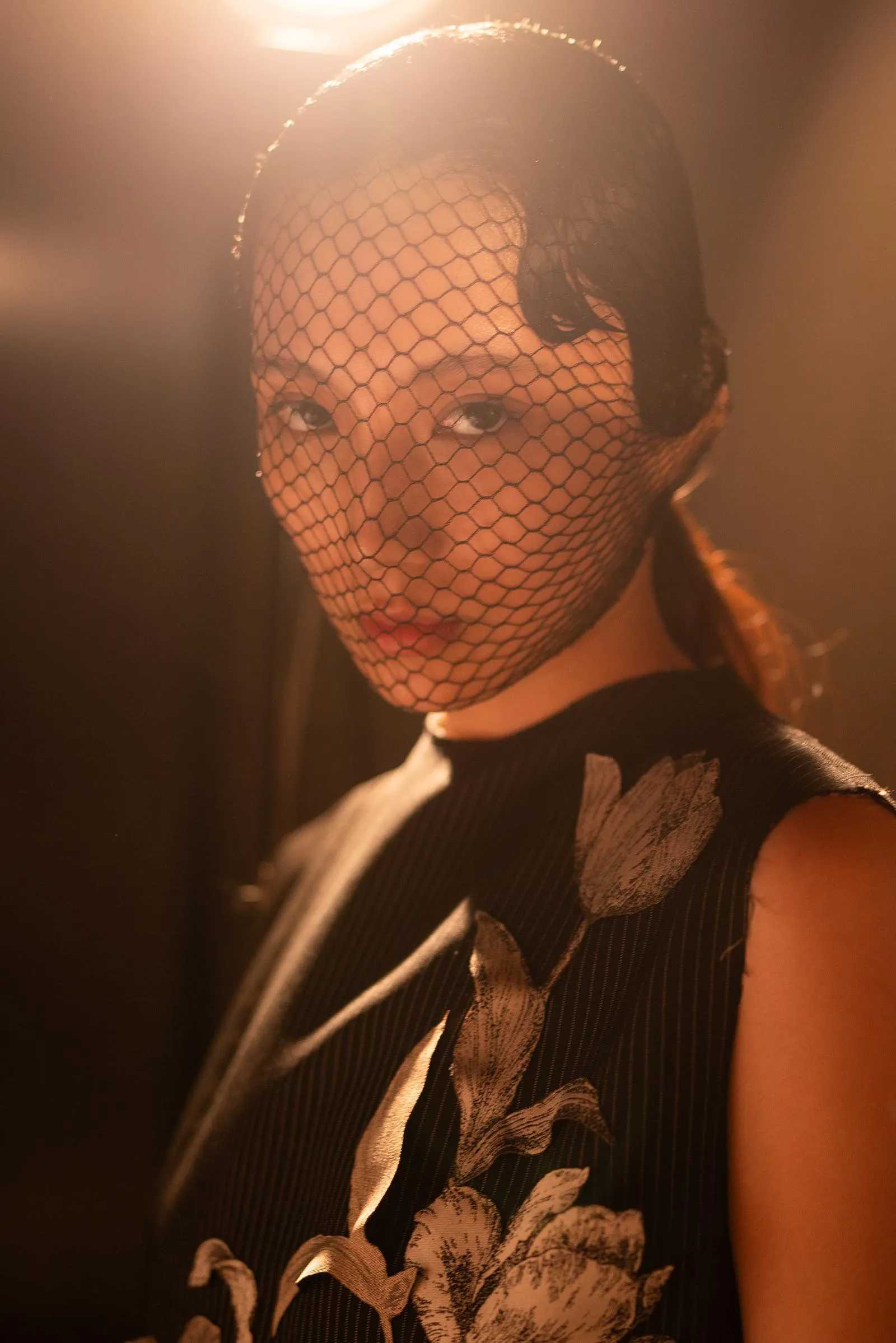 Person wearing a hexagonal fishnet veil over their face with bold makeup and a floral-patterned top in warm backlight.