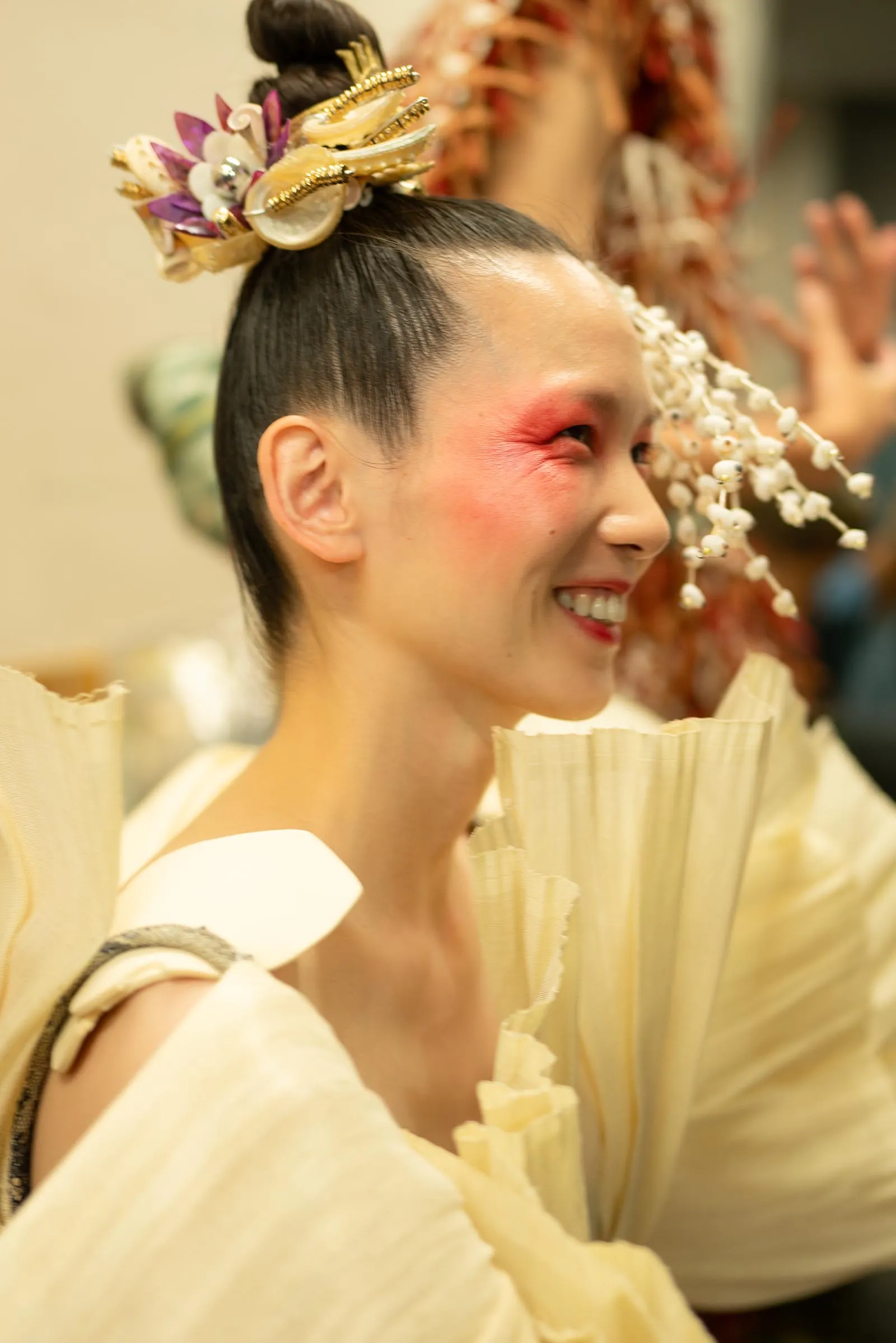 Smiling person in traditional attire with an elaborate gold and flower hair ornament, white makeup, and pink blush seen in profile.