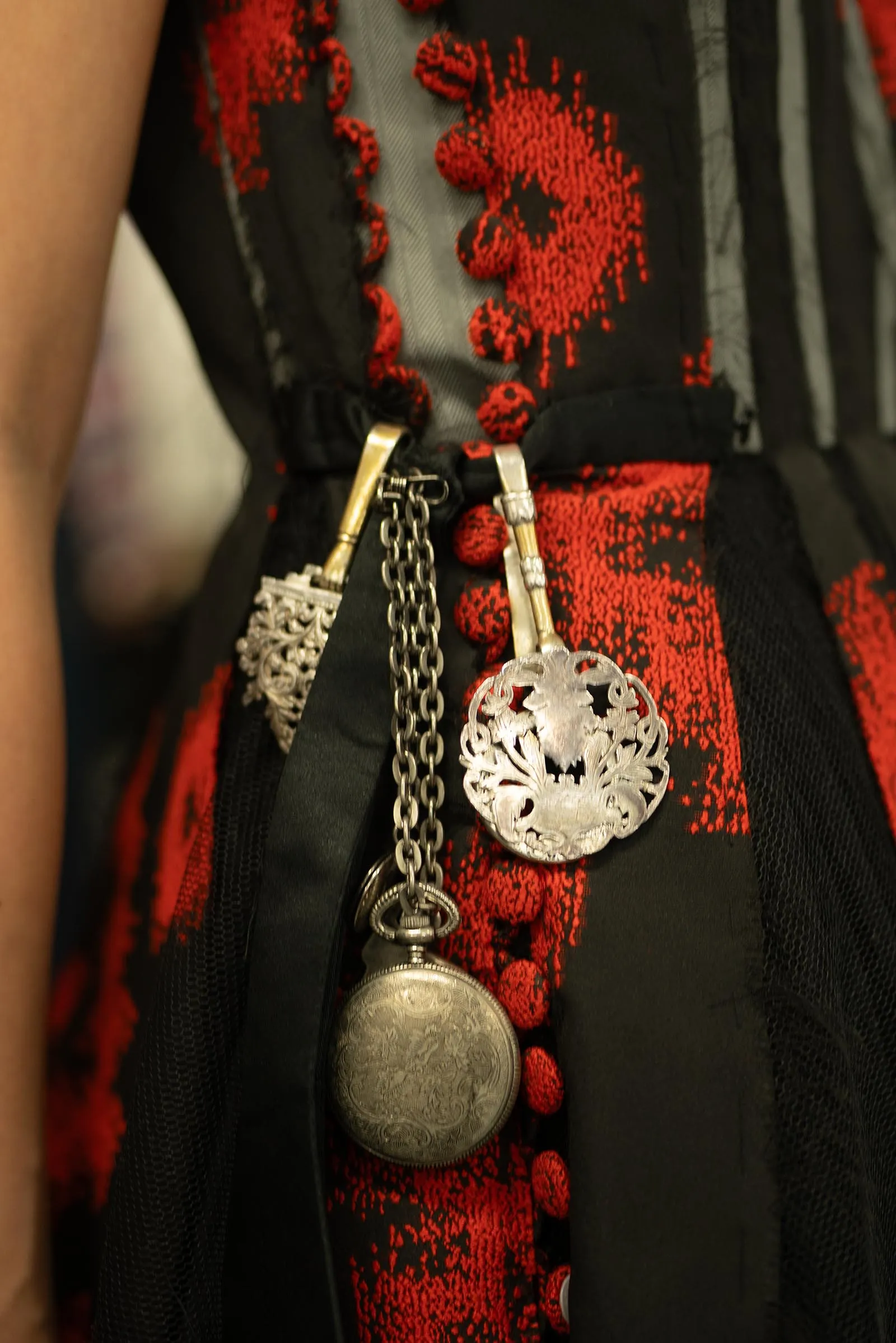 Close-up of a person wearing a red and black patterned dress with metallic necklace pendants and chains hanging at the chest.