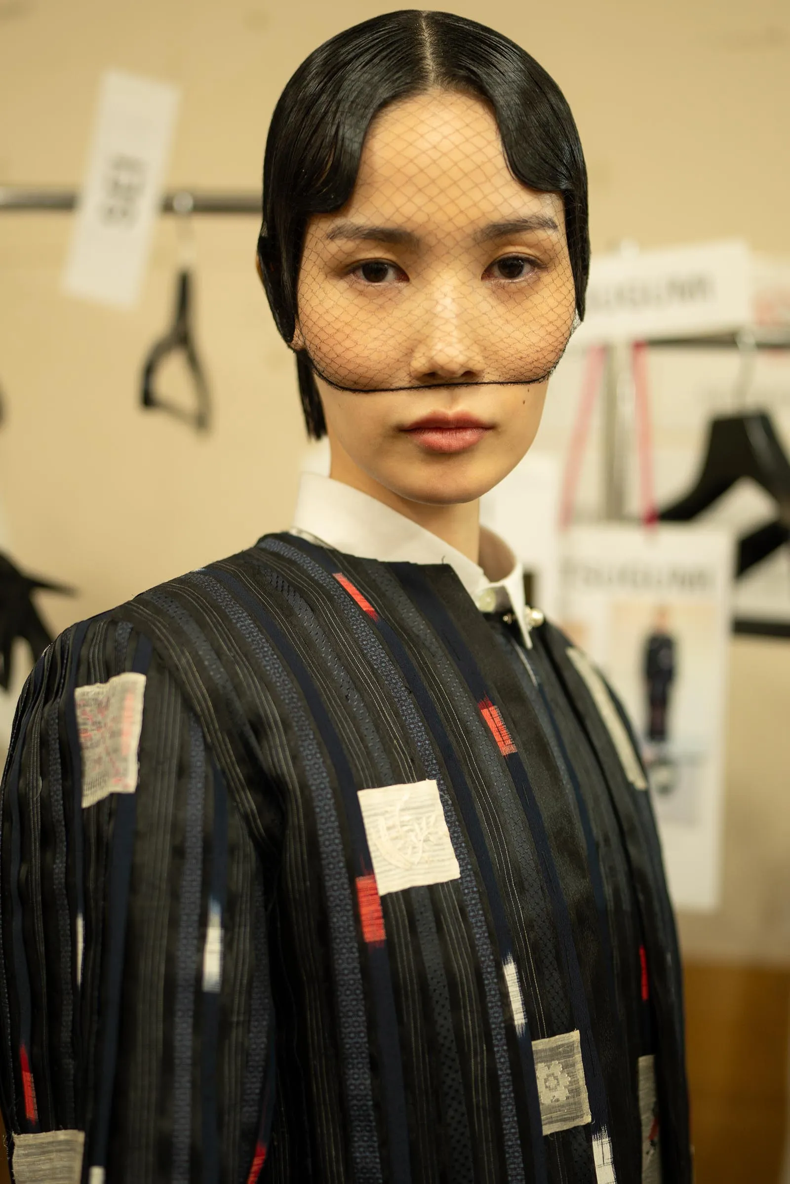 Model wearing a dark pinstriped outfit with fabric patch pockets and a mesh veil across the face in a fashion studio setting.