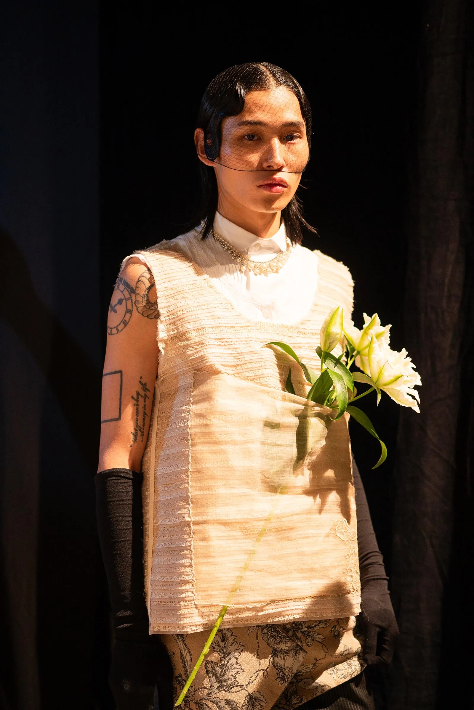Person on a dim stage wearing a cream textured dress, veil over face, necklace, and holding a white lily bouquet.
