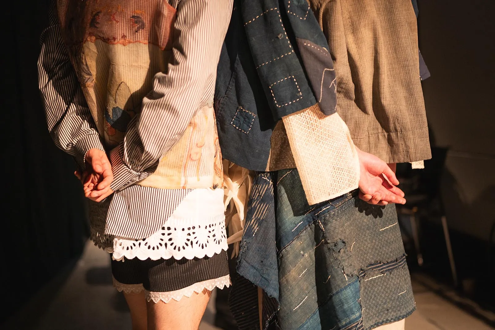 Two people wearing layered patchwork clothing, standing back-to-back; visible fabrics include striped shirts, denim patches, and a white lace hem.