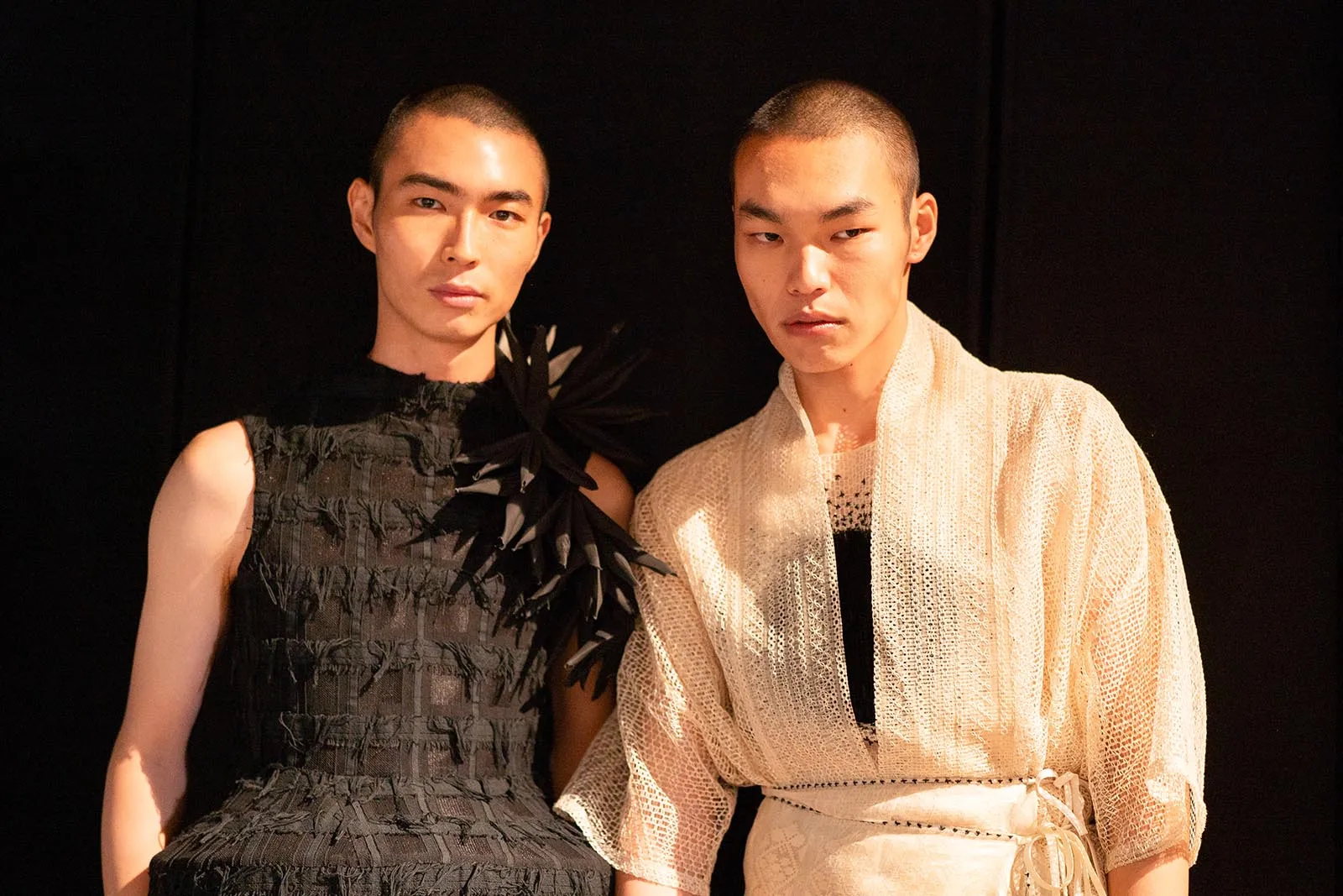 Two fashion models pose side by side in dark, avant-garde outfits: one in a black textured dress with feather accents, the other in a white lace robe over a black top.