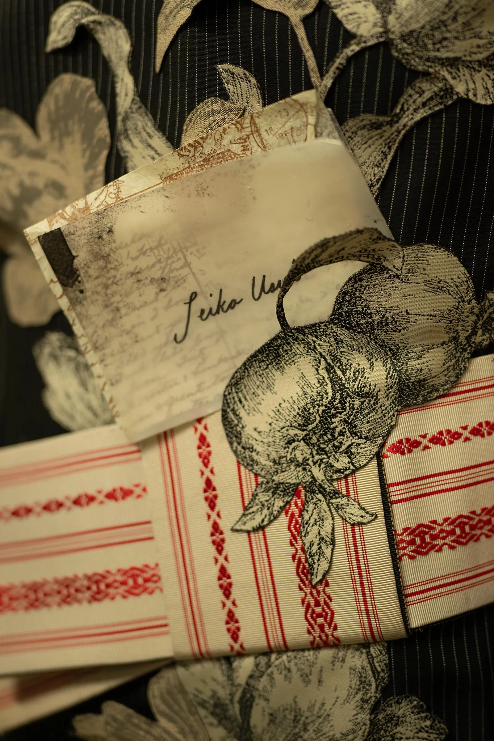Close-up of beige fabric with red embroidery and a folded vintage-looking card with cursive handwriting resting on a dark, patterned background.