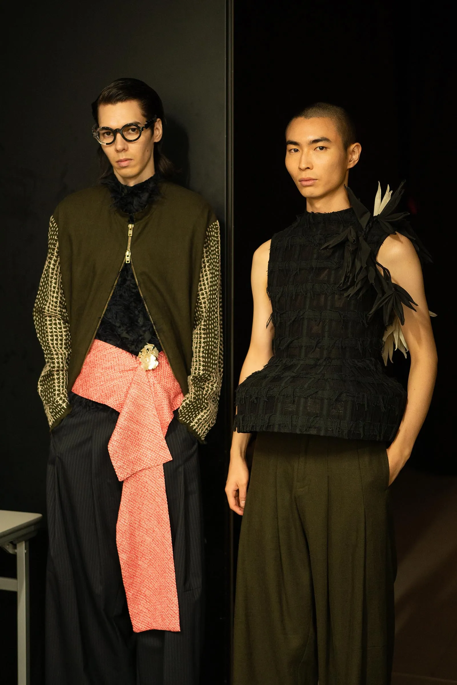 Two models pose in dark fashion outfits: one in olive jacket, net sleeves, pink sash; the other in black textured top with feather details and olive trousers.